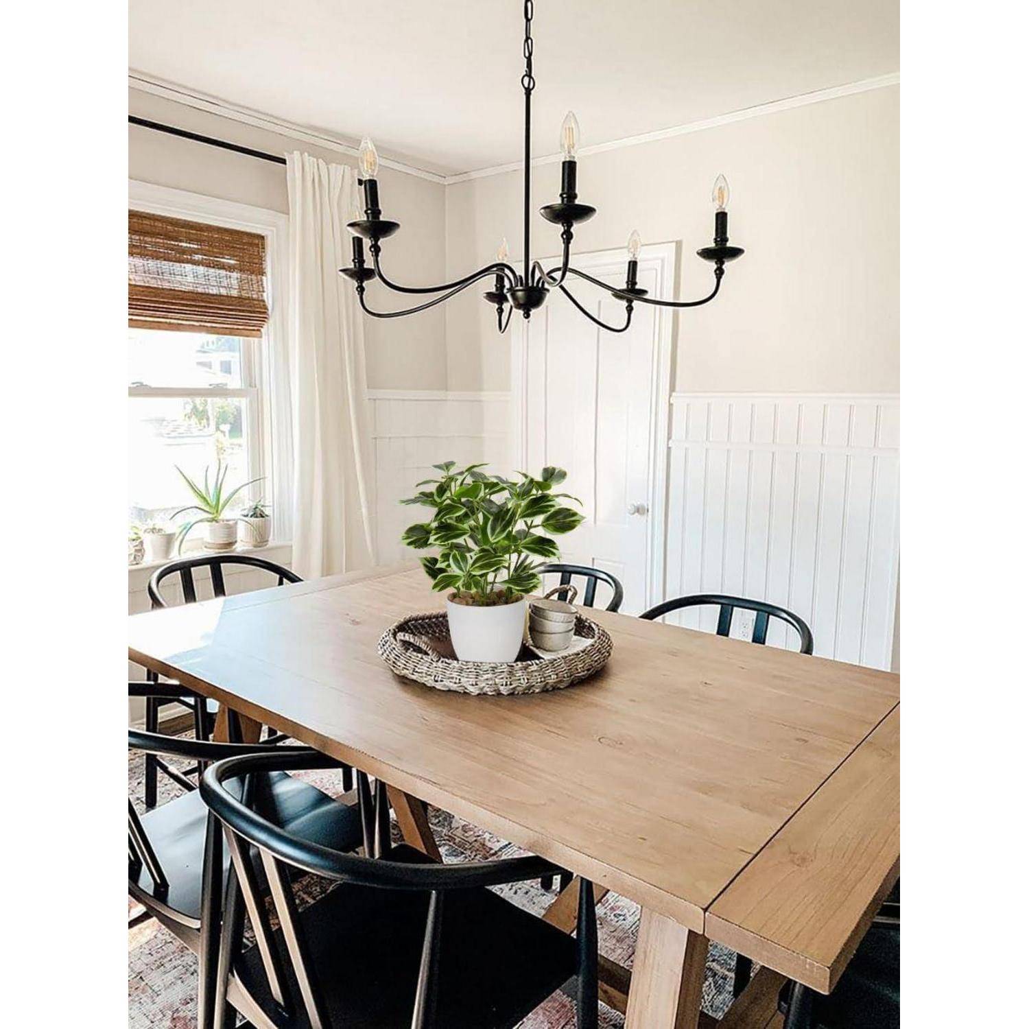 Ein Esszimmer mit einem Holztisch, schwarzen Stühlen und einer zentralen Pflanze in einem weißen Topf. Ein schwarzer Kronleuchter hängt darüber, und ein Fenster mit Jalousien ist zu sehen.