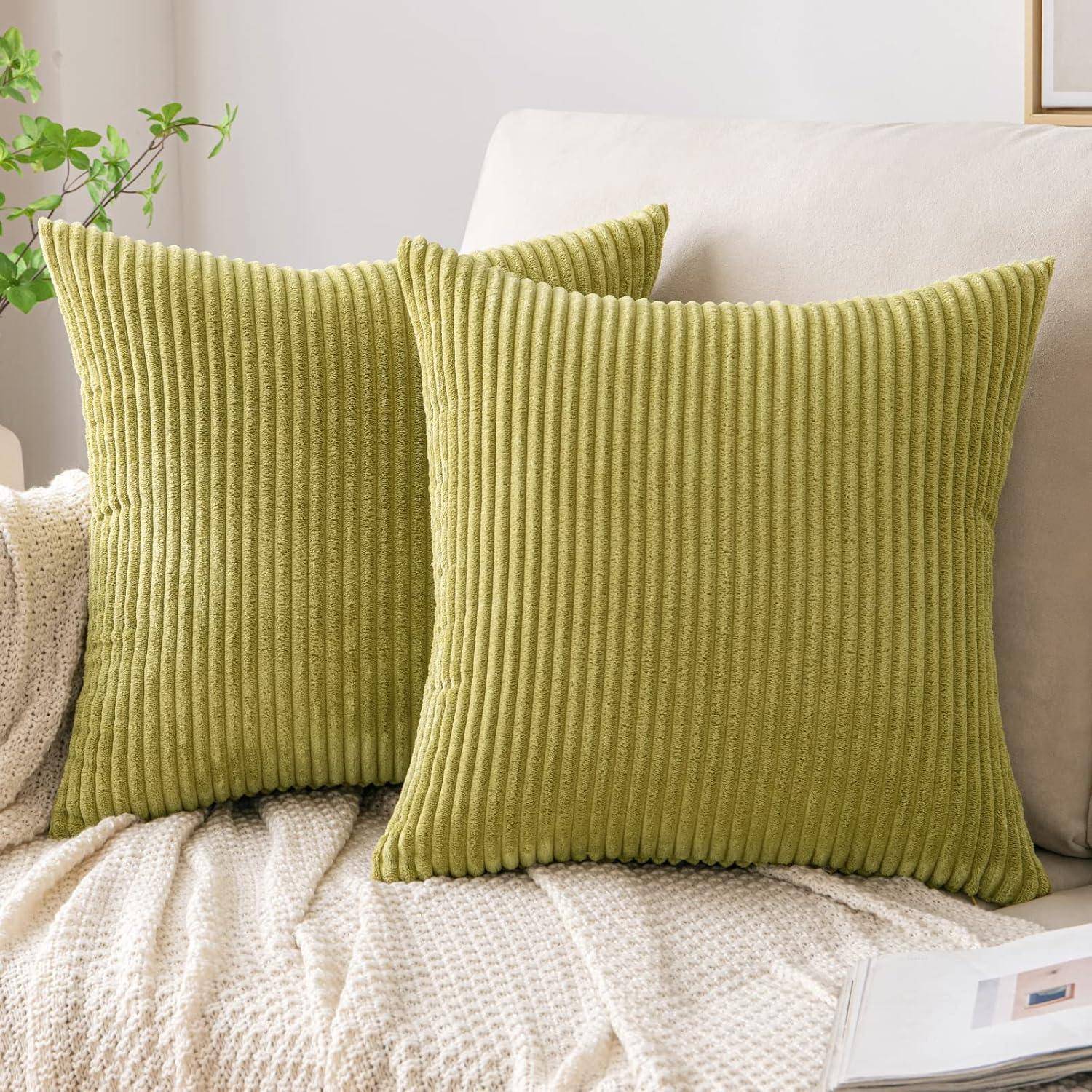 Zwei grüne Cordkissen auf einer beigen strukturierten Decke, auf einem hellen Sofa platziert, mit einer Pflanze in der Ecke, die eine gemütliche Atmosphäre schafft.