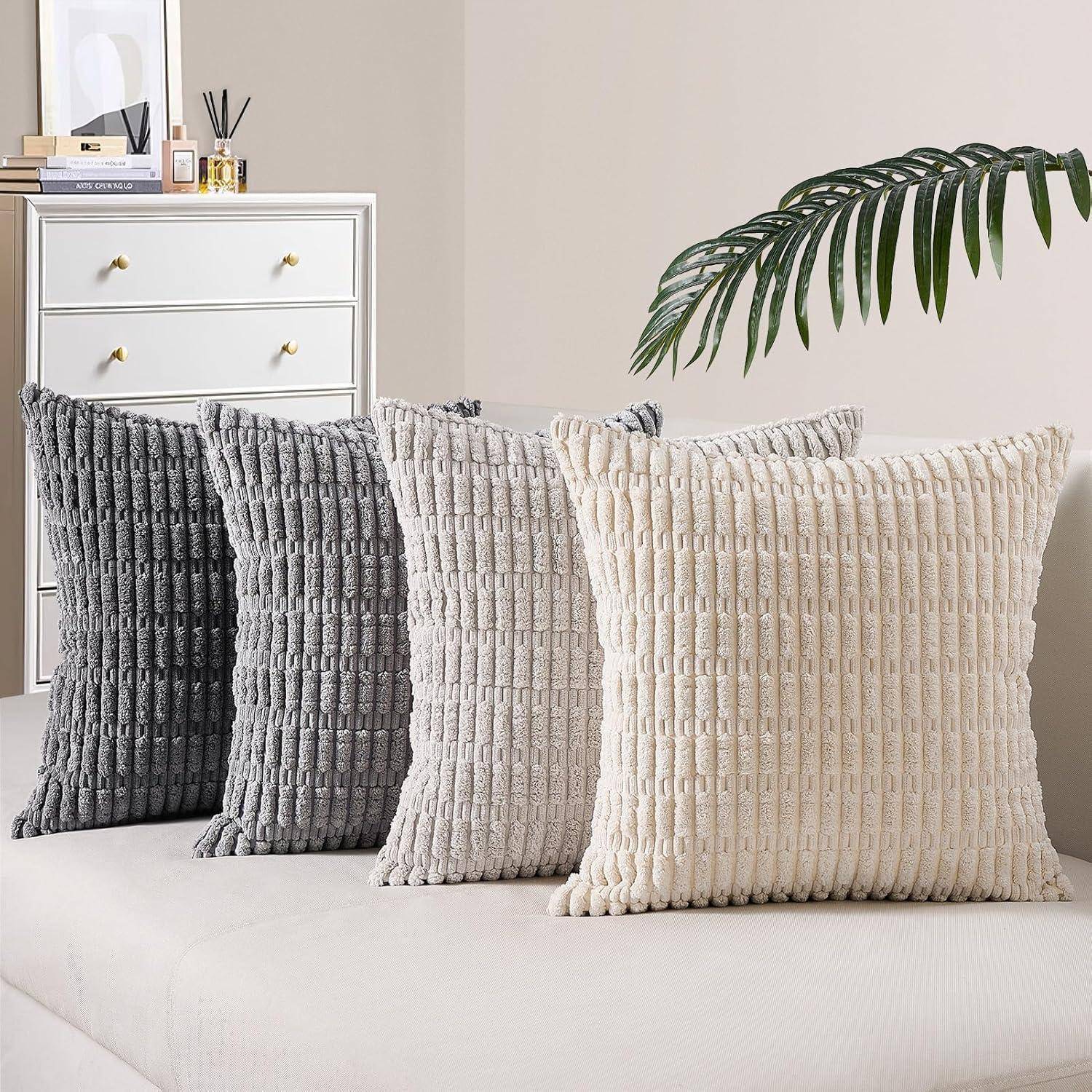 Vier strukturierte Kissen in Grau- und Beigetönen sind auf einem modernen Sofa angeordnet, mit einer weißen Kommode und einer Pflanze im Hintergrund.