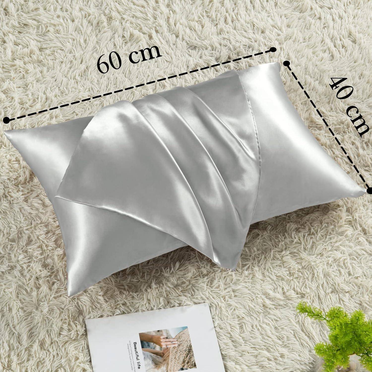 Ein silberner Seidenkissen mit gefalteter Gestaltung liegt auf einer strukturierten Oberfläche. Die Abmessungen sind mit 60 cm x 40 cm gekennzeichnet. Ein Booklet liegt in der Nähe.
