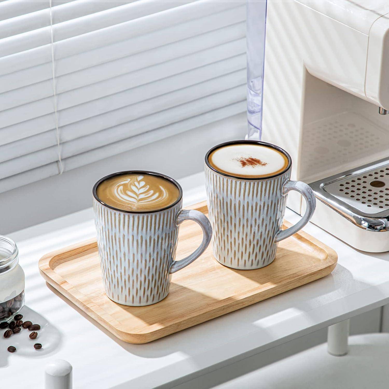 Zwei Keramiktassen auf einem Holztablett neben einer Kaffeemaschine; eine enthält einen Cappuccino mit Latte Art, die andere Milchschaum mit Zimt.