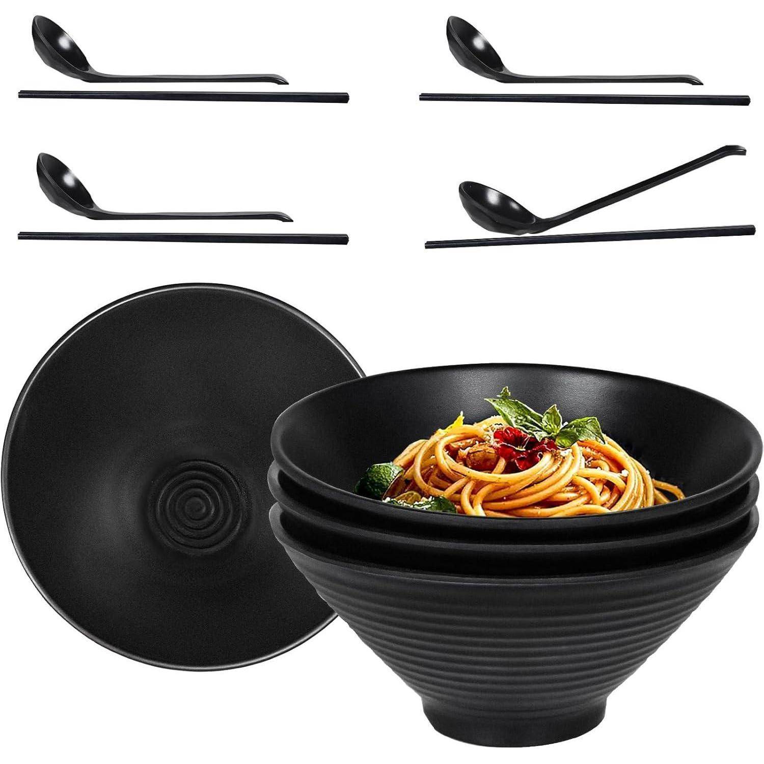 Vier schwarze Schüsseln und vier schwarze Löffel. Eine Schüssel enthält Spaghetti, garniert mit Kräutern und Tomaten.