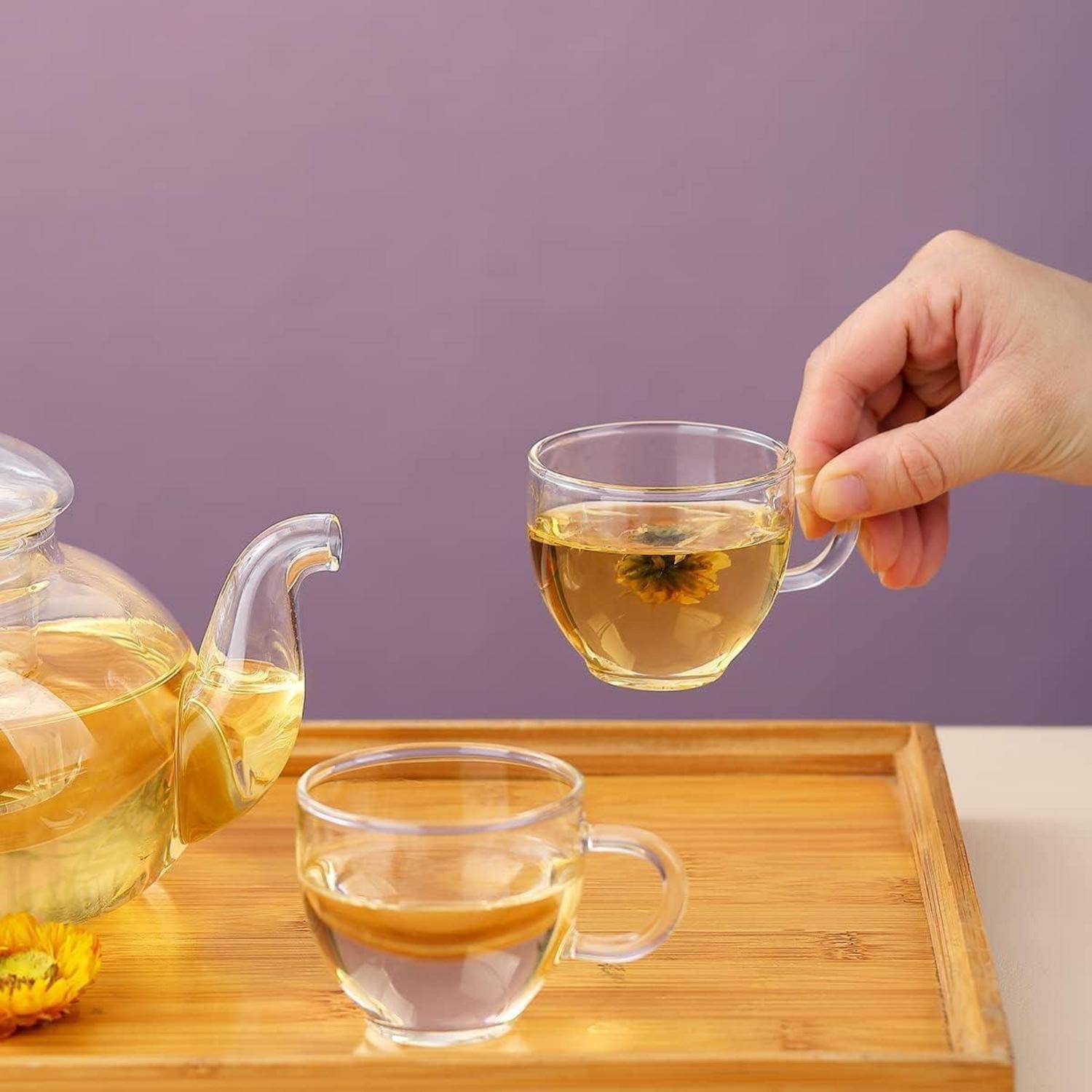 Eine Hand, die eine durchsichtige Glasteekanne mit blühender Blumentee hält, neben einer Teekanne und einer weiteren gefüllten Tasse auf einem Holztablett vor einem lila Hintergrund.