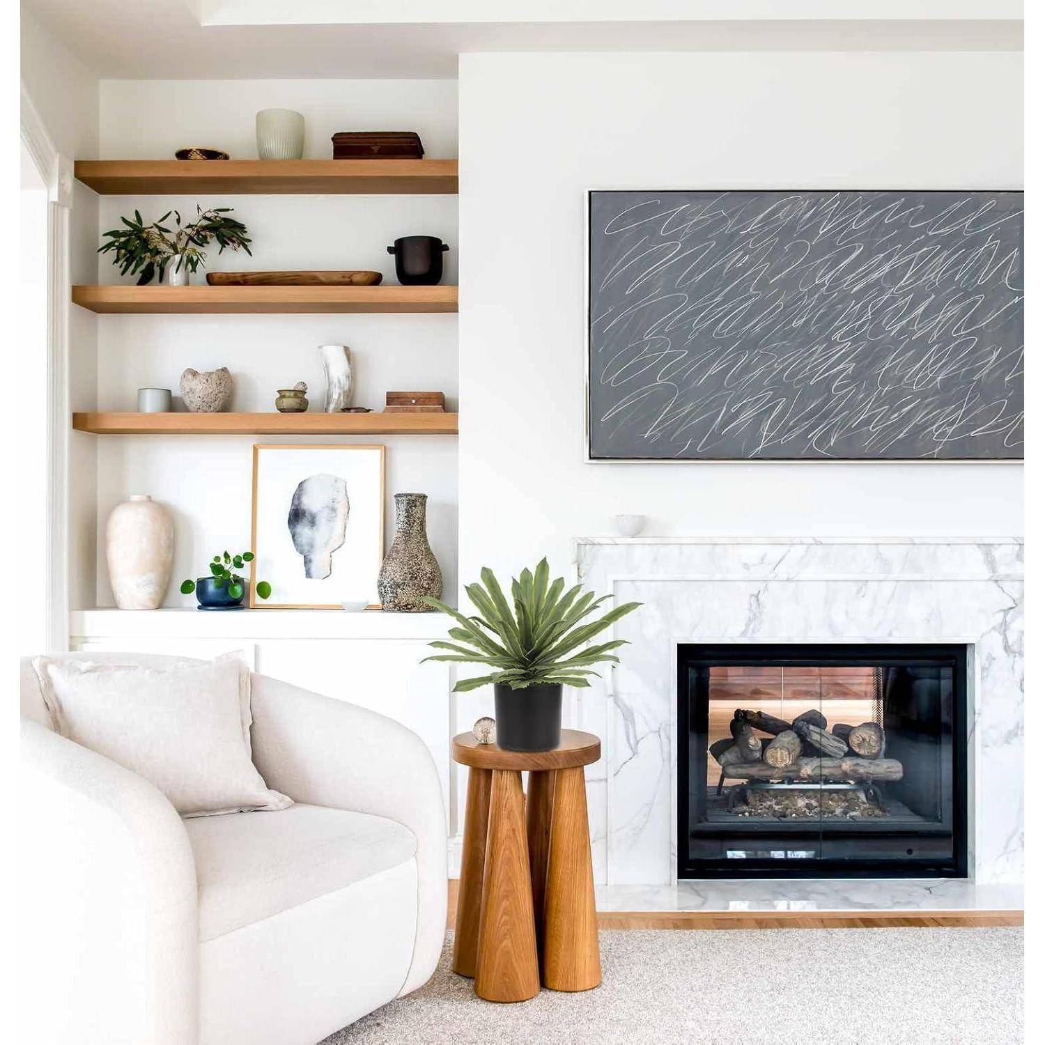 Ein modernes Wohnzimmer mit einem weißen Sessel, minimalistischen Holzregalen und einem Marmorkamin. Über dem Kamin befindet sich abstrakte Wandkunst.