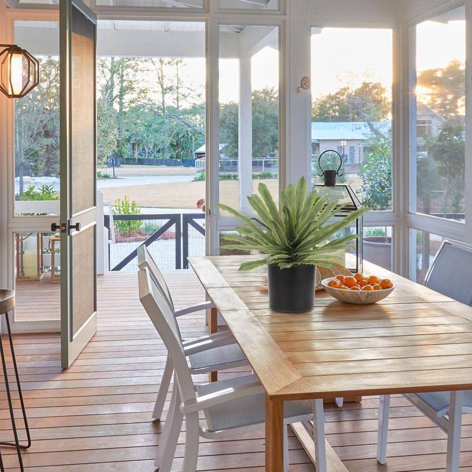 Eine sonnendurchflutete, überdachte Veranda mit einem Holztisch, einer Topfpflanze und einer Schüssel mit Orangen. Weiße Stühle umgeben den Tisch, und von draußen ist ein Blick in einen Garten zu sehen.