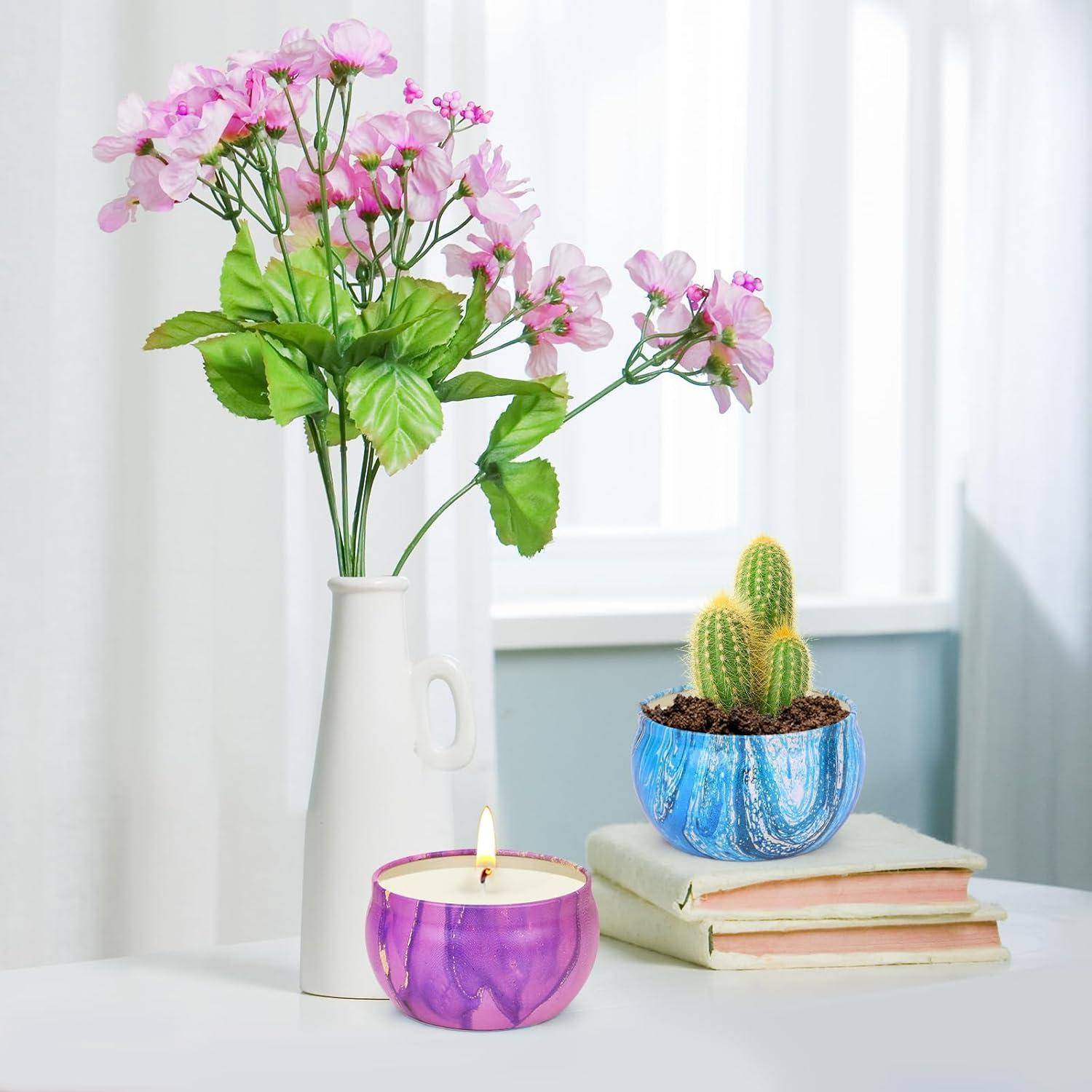 Eine weiße Vase mit rosa Blumen, ein blauer Topf mit einem Kaktus auf gestapelten Büchern und eine brennende lila Kerze auf einem weißen Tisch neben einem Fenster.