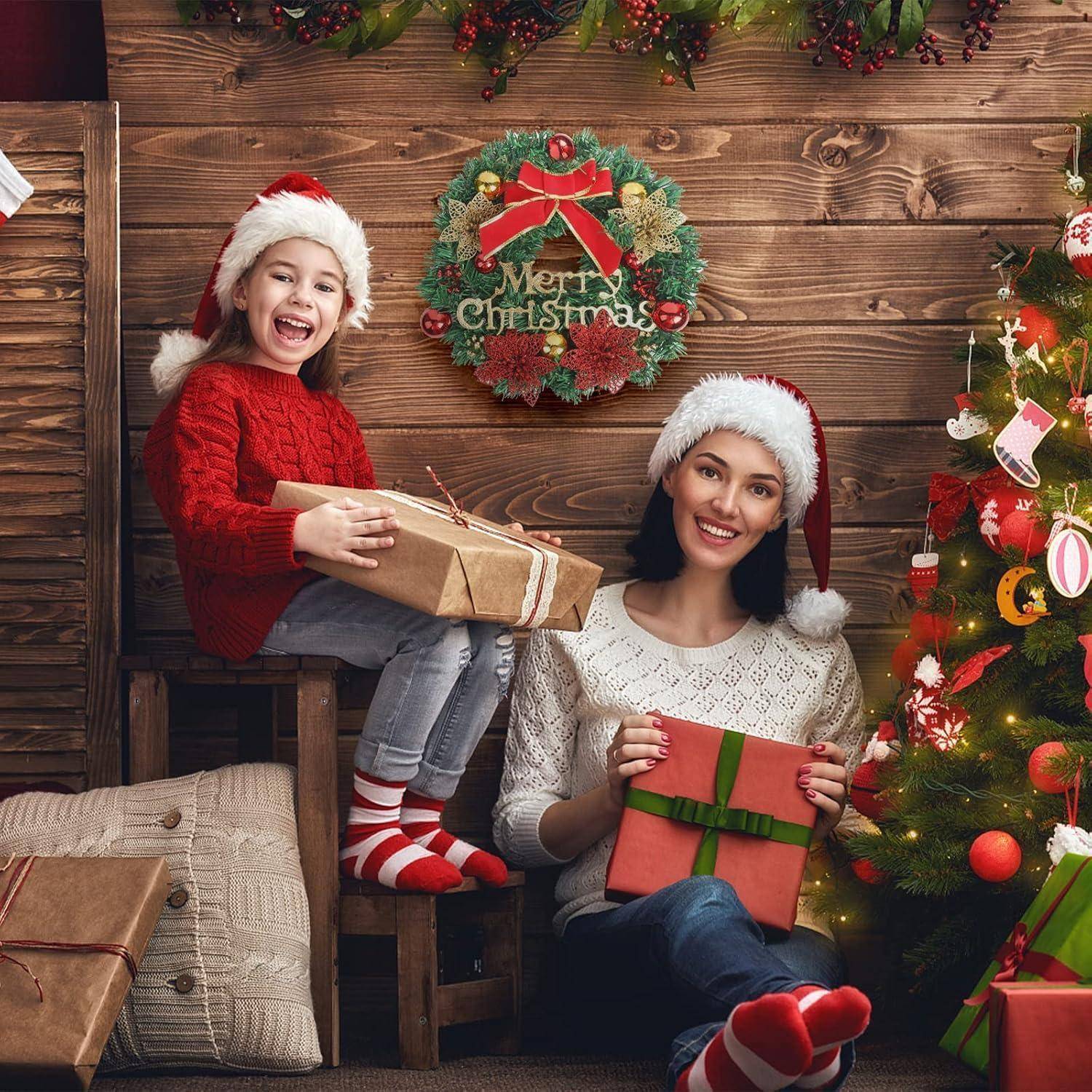 Eine Mutter und ihre Tochter sitzen neben einem Weihnachtsbaum, beide tragen Weihnachtsmützen und halten verpackte Geschenke. Ein Adventskranz mit der Aufschrift ‚Frohe Weihnachten' hängt im Hintergrund.