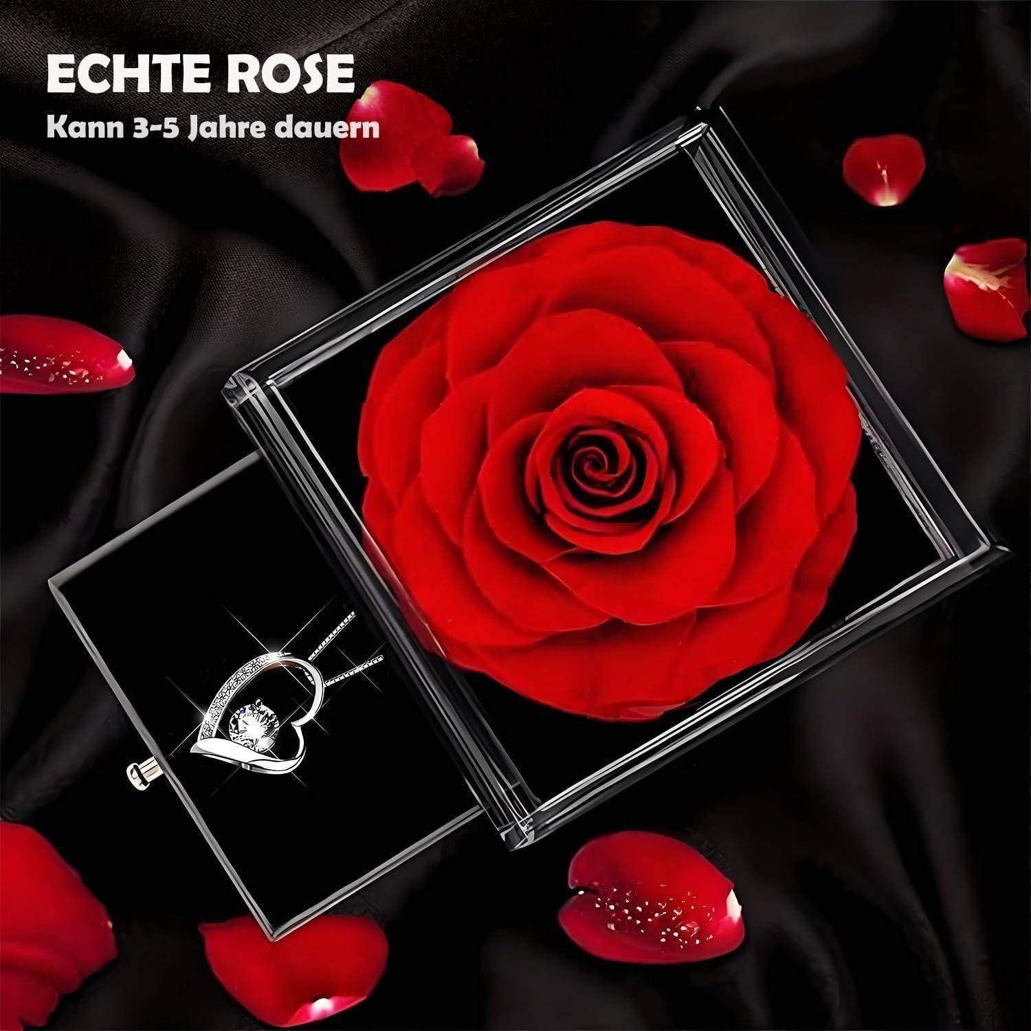 Eine rote Rose in einer durchsichtigen Schatulle liegt neben einer Ringbox mit einem Diamantring, vor einem schwarzen Seidenhintergrund mit Rosenblüten.