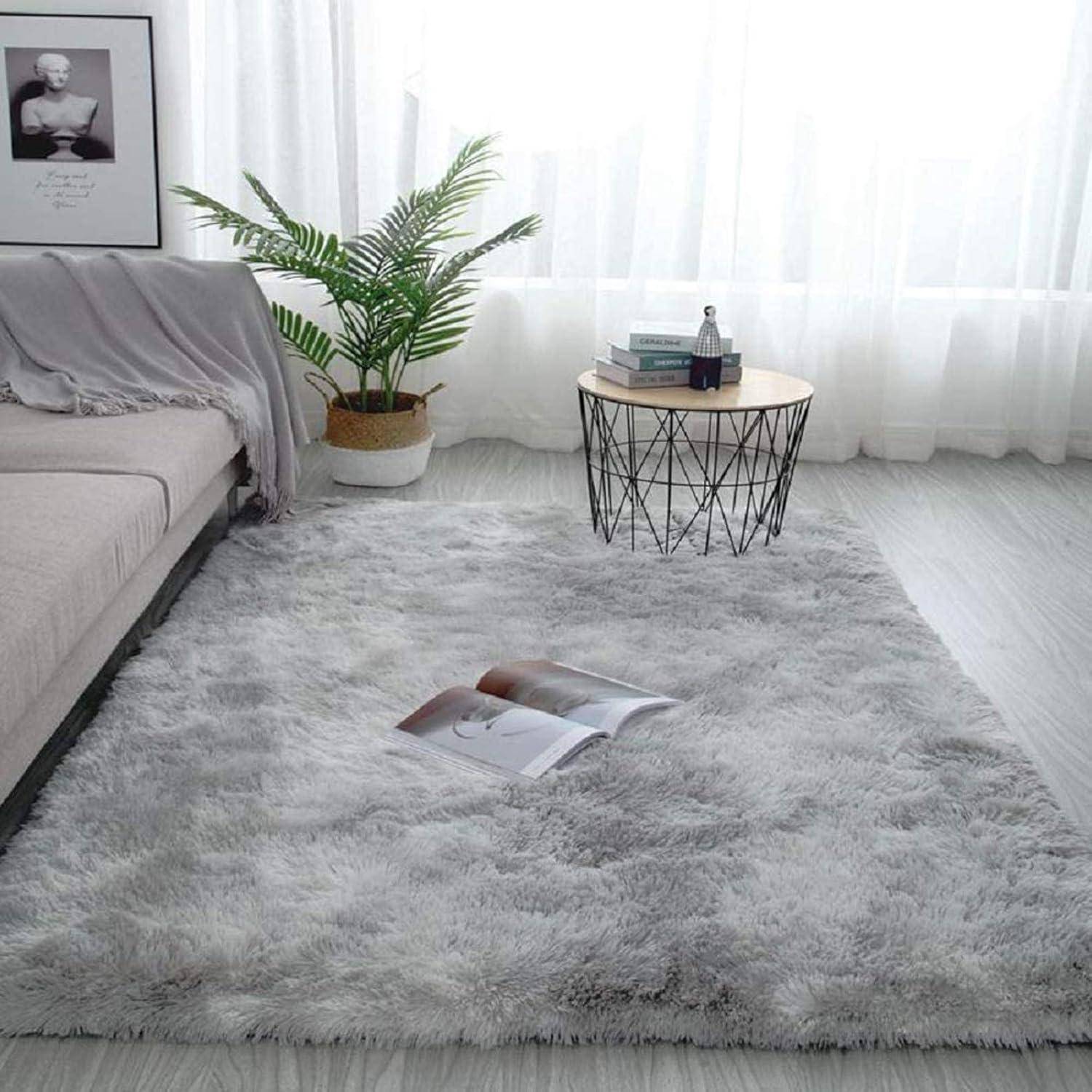Ein gemütliches Wohnzimmer mit einem grauen Shaggy-Teppich, einem hellgrauen Sofa und einem runden weißen Tisch, auf dem Bücher und eine kleine Vase stehen, nahe großen Fenstern.