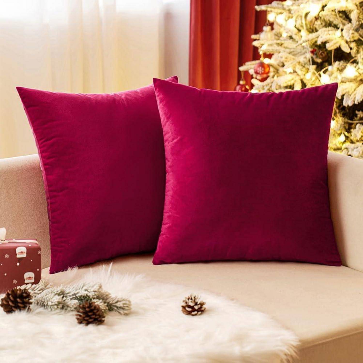 Zwei rote Kissen auf einem beigen Sofa neben einem geschmückten Weihnachtsbaum mit einem roten Vorhang im Hintergrund.