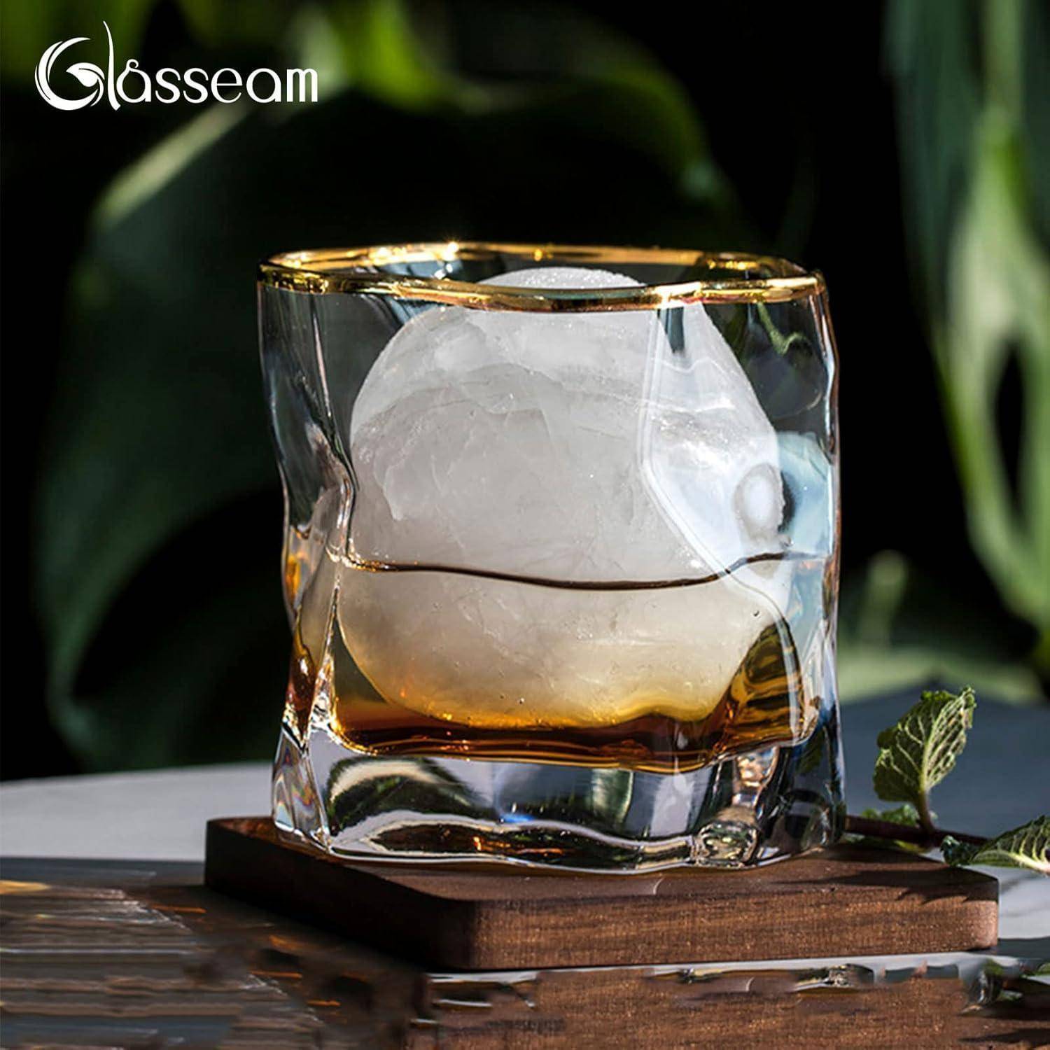 Ein klares Glas mit einer einzigartigen, wellenförmigen Gestalt hält eine runde Eiskugel auf einem Untersetzer, vor einem verschwommenen dunklen Hintergrund mit Grünpflanzen.