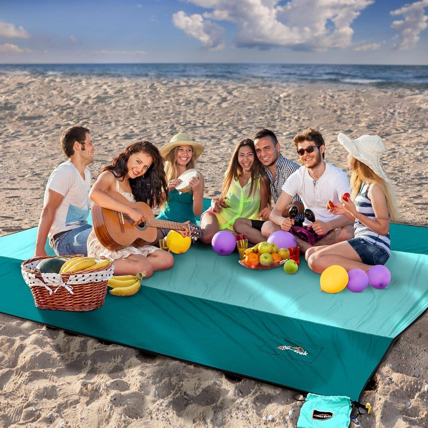Eine Gruppe von Menschen sitzt auf einer großen Decke am Strand. Eine Person spielt Gitarre, während andere Snacks und Getränke genießen. Ein heller, sonniger Tag.