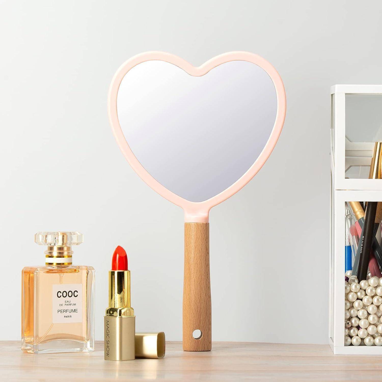 Ein Spiegel mit Holzgriff in Herzform steht auf einem Regal neben einer Flasche Parfüm, einem Lippenstift und einem durchsichtigen Behälter mit Make-up-Pinseln.