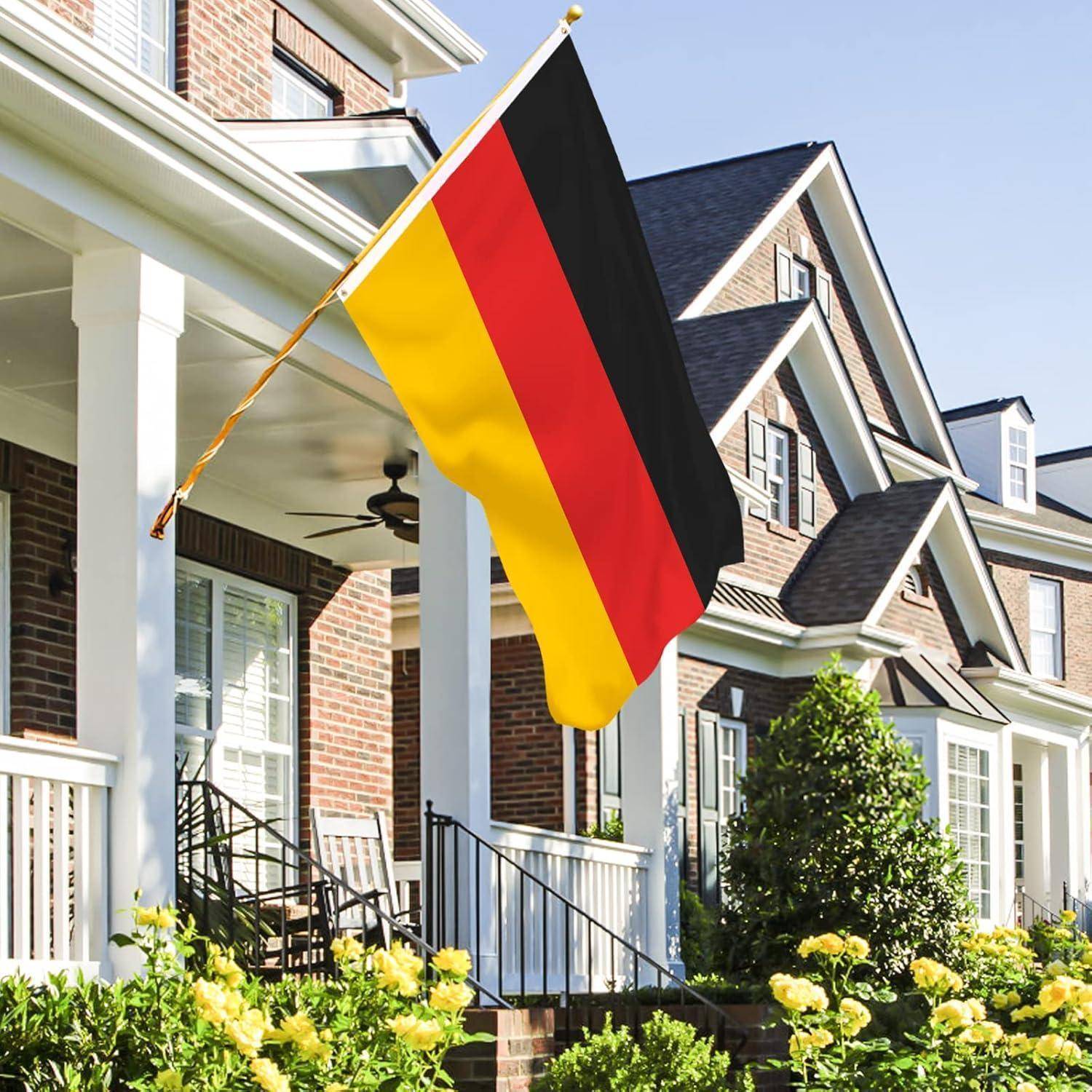 Die deutsche Flagge weht auf der Veranda eines Vorstadthauses mit einem gepflegten Garten und leuchtendgelben Blumen im Vordergrund.