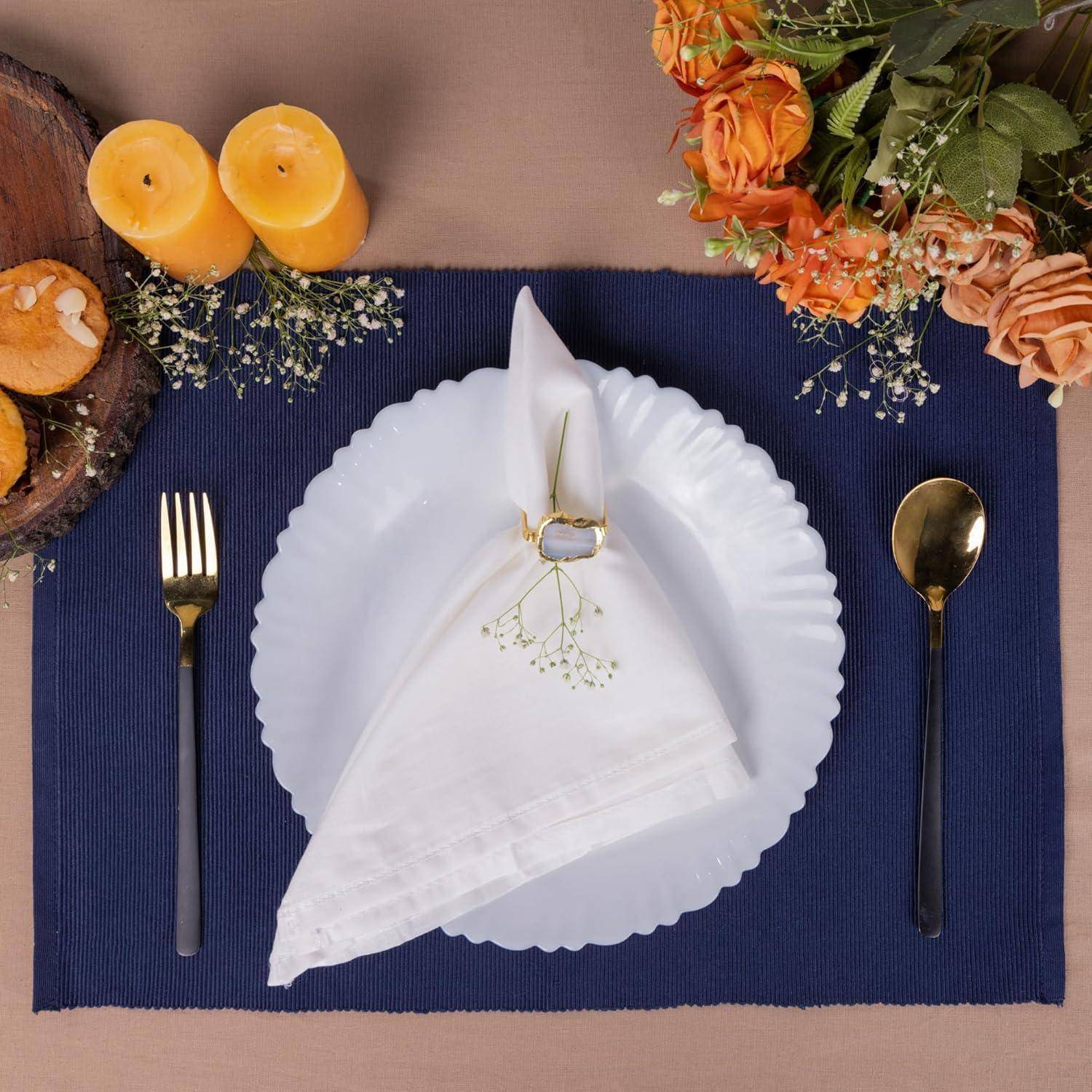 Ein sorgfältig gedeckter Tisch mit einem weißen Teller, goldenen Bestecken, einer weißen Serviette mit einem Serviettenring und dekorativen orangefarbenen Blumen und Kerzen.