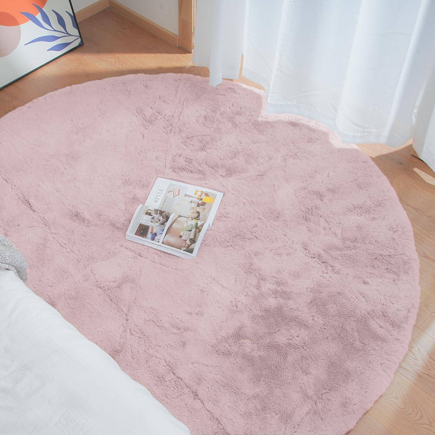 Ein kleiner, gemütlicher Raum mit einem runden, rosa Teppich auf einem hellen Holzboden, neben einem Bett. Eine Zeitschrift liegt auf dem Teppich in der Nähe von weichen, weißen Vorhängen.
