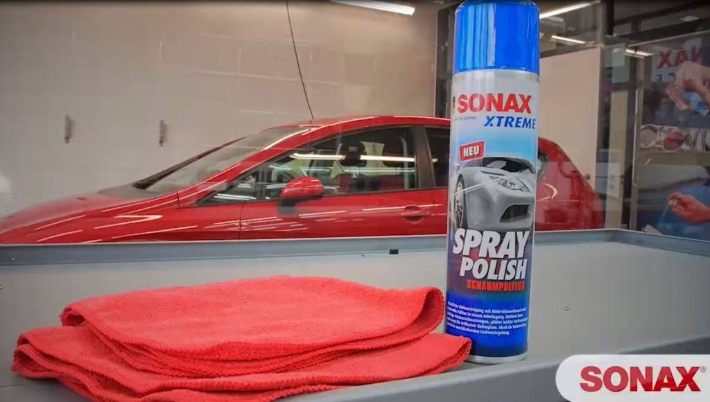 Eine Dose "SONAX Xtreme Spraypolitur"" und ein rotes Tuch auf einer Oberfläche, mit einem roten Auto im Hintergrund, was Autopflegeprodukte andeutet.