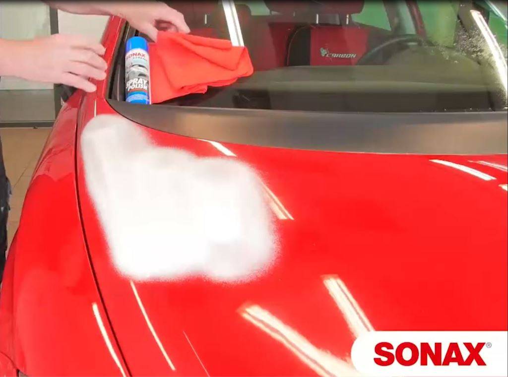 Eine Person poliert eine rote Motorhaube eines Autos mit einem orangefarbenen Tuch und Autopolitur, wobei Lichtreflexe auf der Oberfläche sichtbar sind.