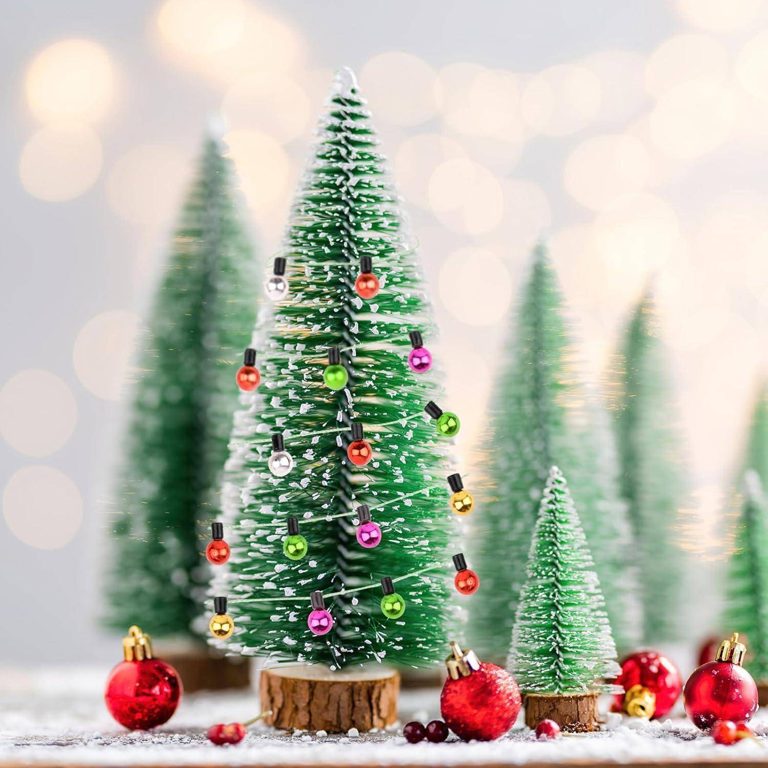 Ein kleiner Weihnachtsbaum, geschmückt mit bunten Ornamenten und Lichtern, umgeben von roten Weihnachtskugeln auf einer Holzbasis mit verschwommenen Lichtern im Hintergrund.