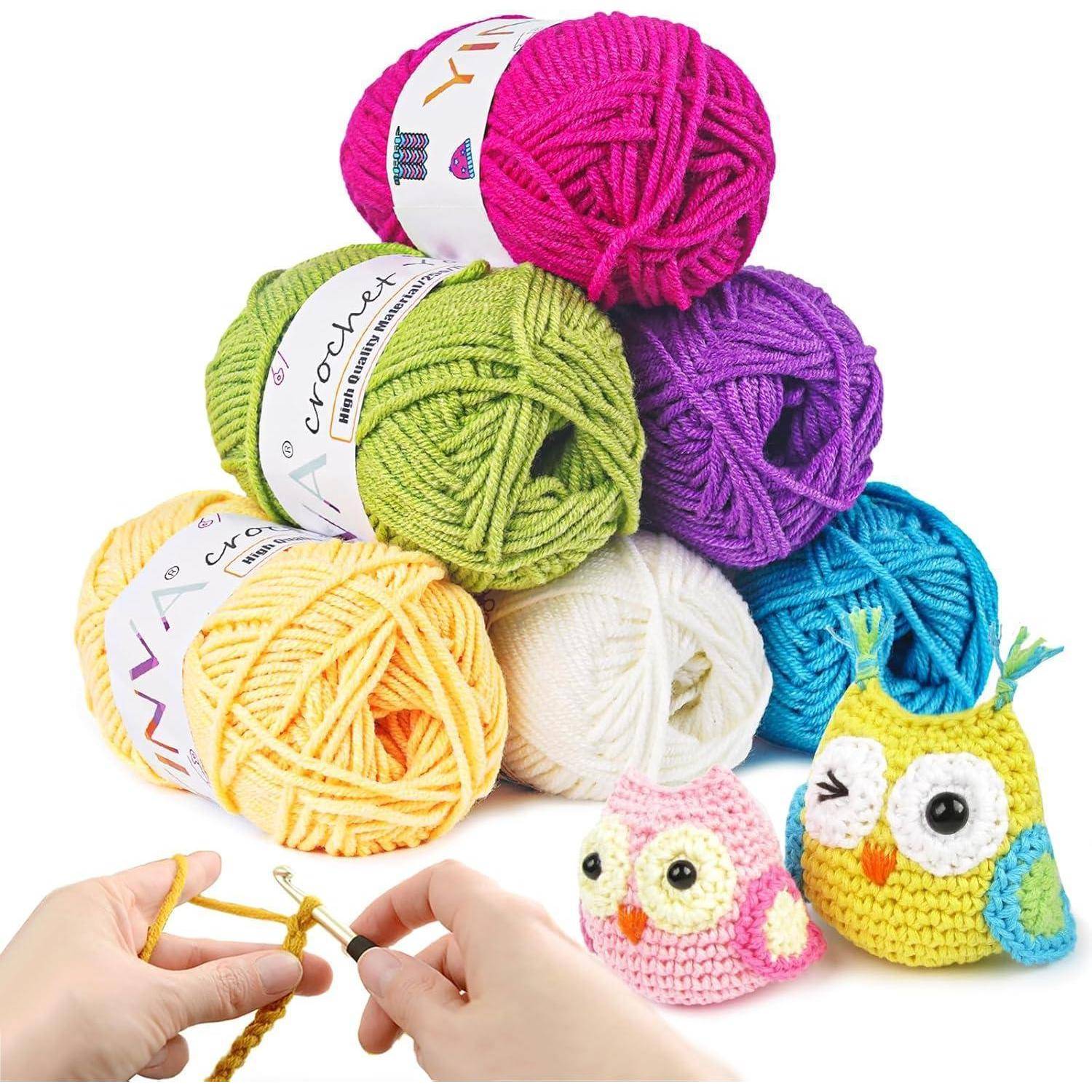 Eine Sammlung bunter Wollbälle, gestapelt mit zwei gehäkelten Eulen; Hände, die mit einer Häkelnadel im Vordergrund häkeln.