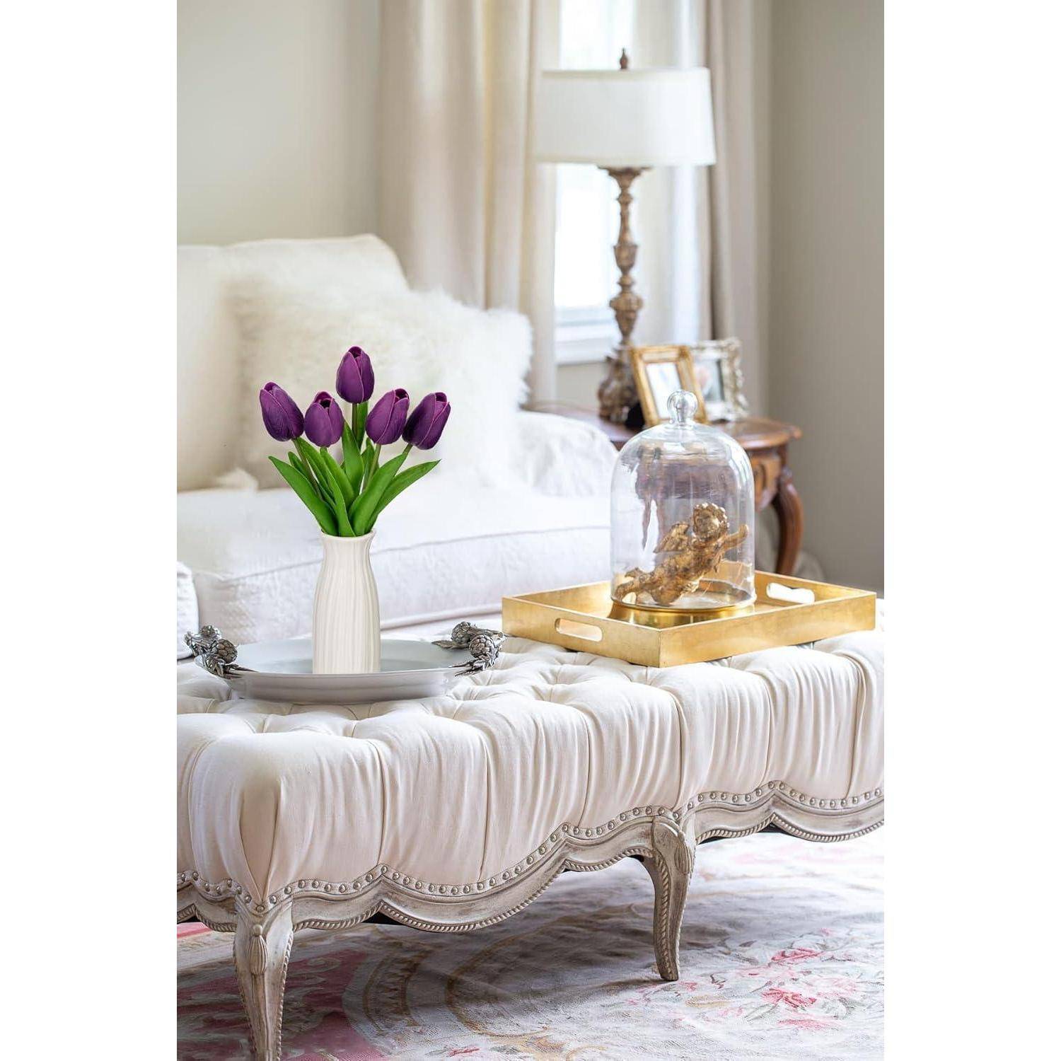Ein Wohnzimmer mit einem weichen, weißen Hocker, einem goldenen Tablett mit Dekoration und einer Vase mit lila Tulpen. Eine Lampe und ein Sessel befinden sich im Hintergrund.