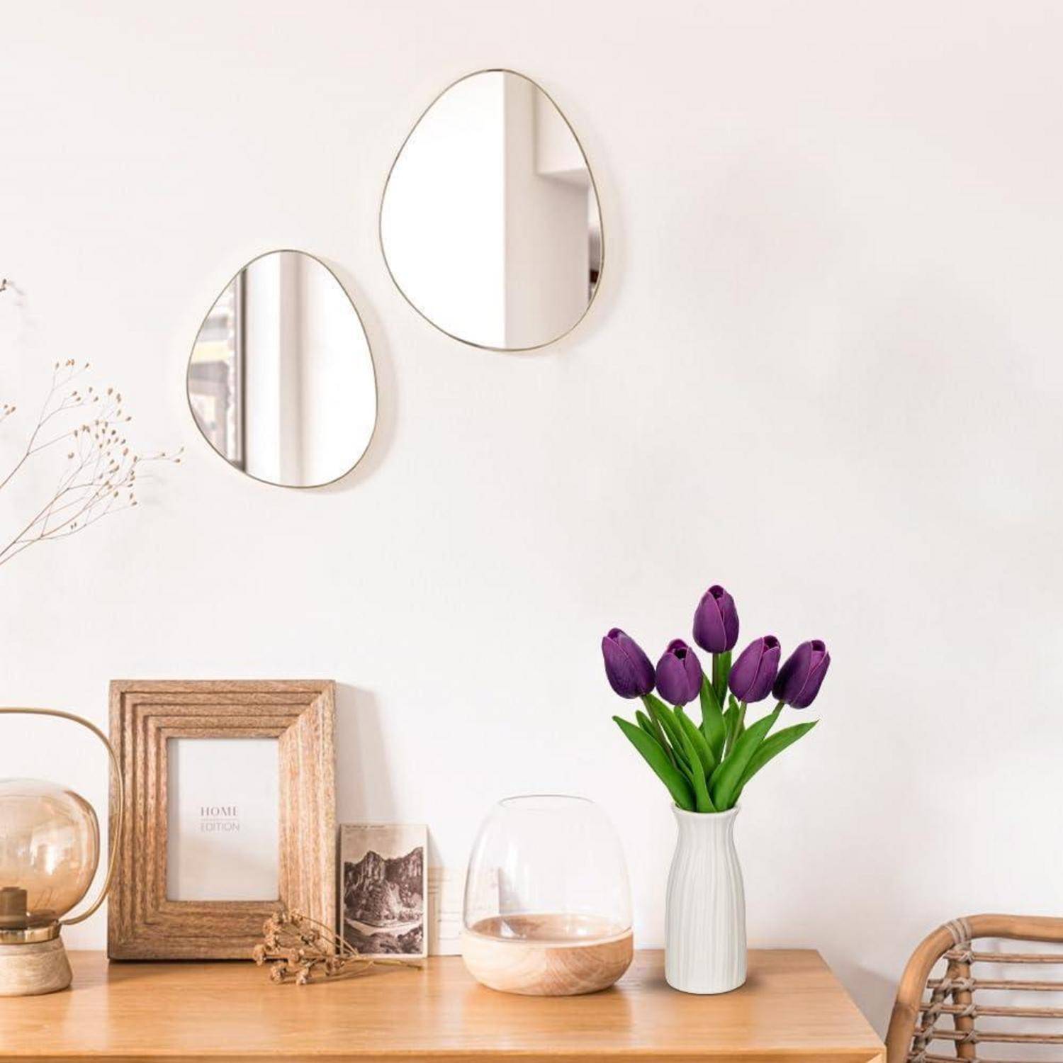 Ein Holztisch mit Dekoration, darunter ein Fotorahmen, Kerzenhalter und eine Vase mit lila Tulpen; zwei ovale Spiegel an der weißen Wand.