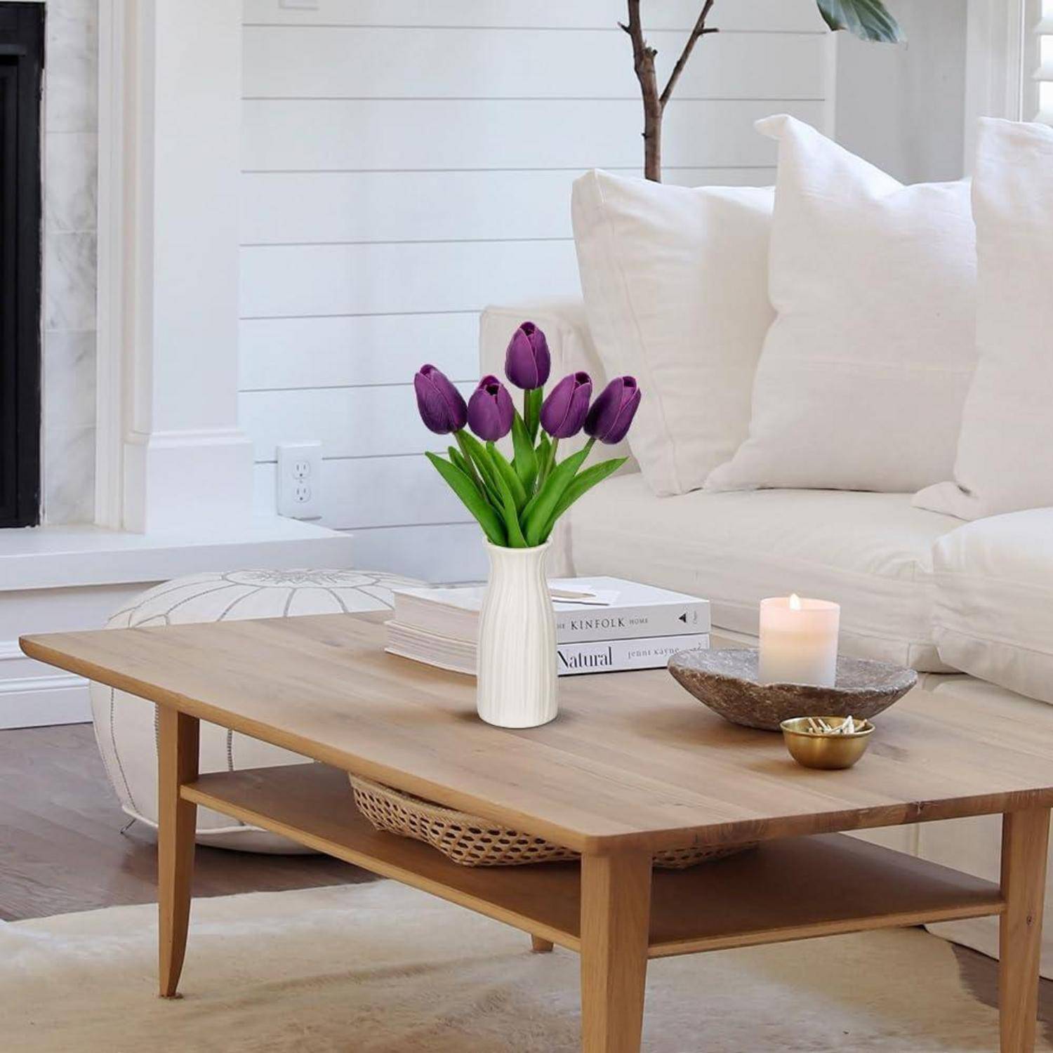 Eine gemütliche Wohnzimmer-Szene mit einem Holzcouchtisch, einer Vase mit lila Tulpen, einer brennenden Kerze und einem Sofa mit weißen Kissen.