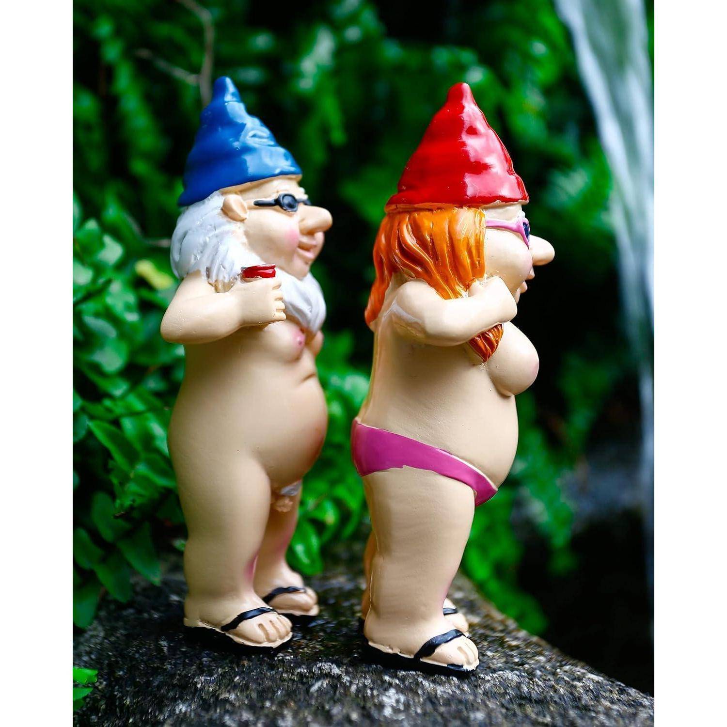 Zwei Gartenzwerge mit blauen und roten Hüten stehen Rücken an Rücken auf einem Steinvorsprung, halten Getränke und sind von üppigem Grün umgeben.