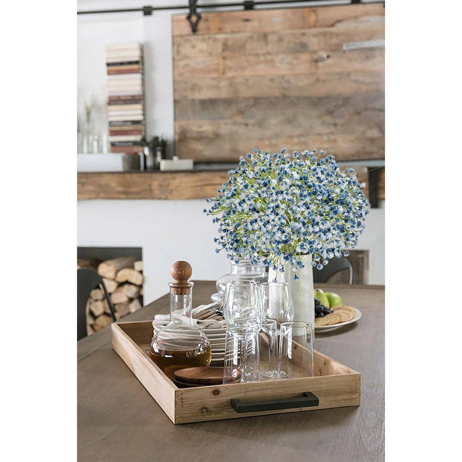 Ein Holztablett auf einem Esstisch trägt Gläser und zwei Karaffen; eine davon mit einem Korken. Ein Blumenstrauß aus kleinen blauen und weißen Blüten ist in der Nähe platziert.