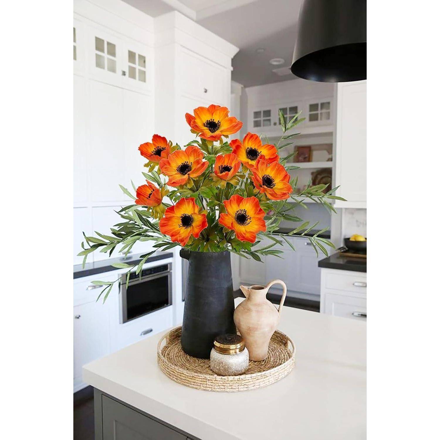 Eine Küchenarbeitsplatte zeigt eine dunkle Vase mit leuchtend orangefarbenen Blumen und grünen Blättern, daneben einen kleinen Krug und ein Gefäß auf einer geflochtenen Matte.