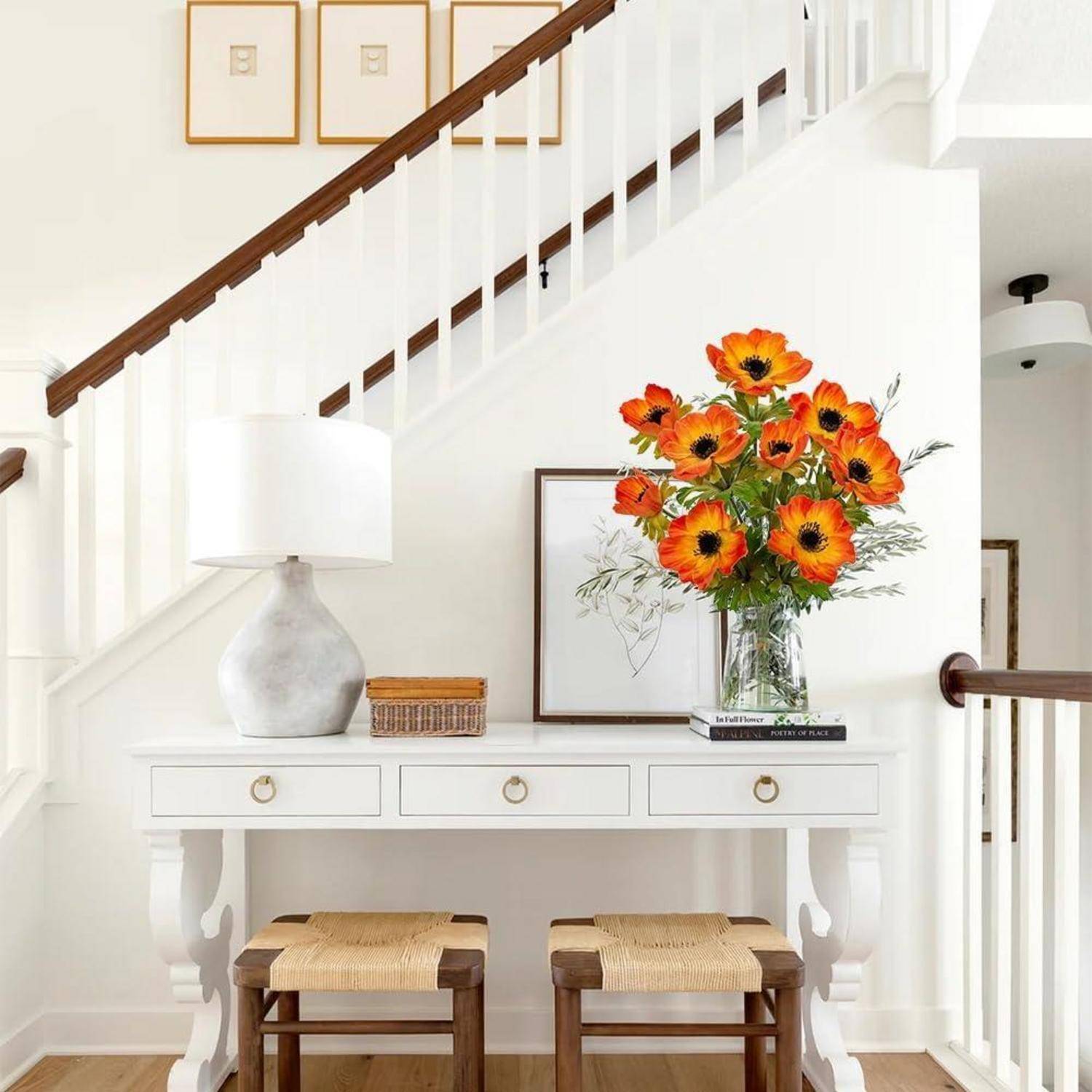 Ein heller Flur mit einem weißen Tisch, orangefarbenen Blumen in einer Vase, zwei Hockern und gerahmter Kunst an der Wand. Eine lichte und luftige Einrichtung.