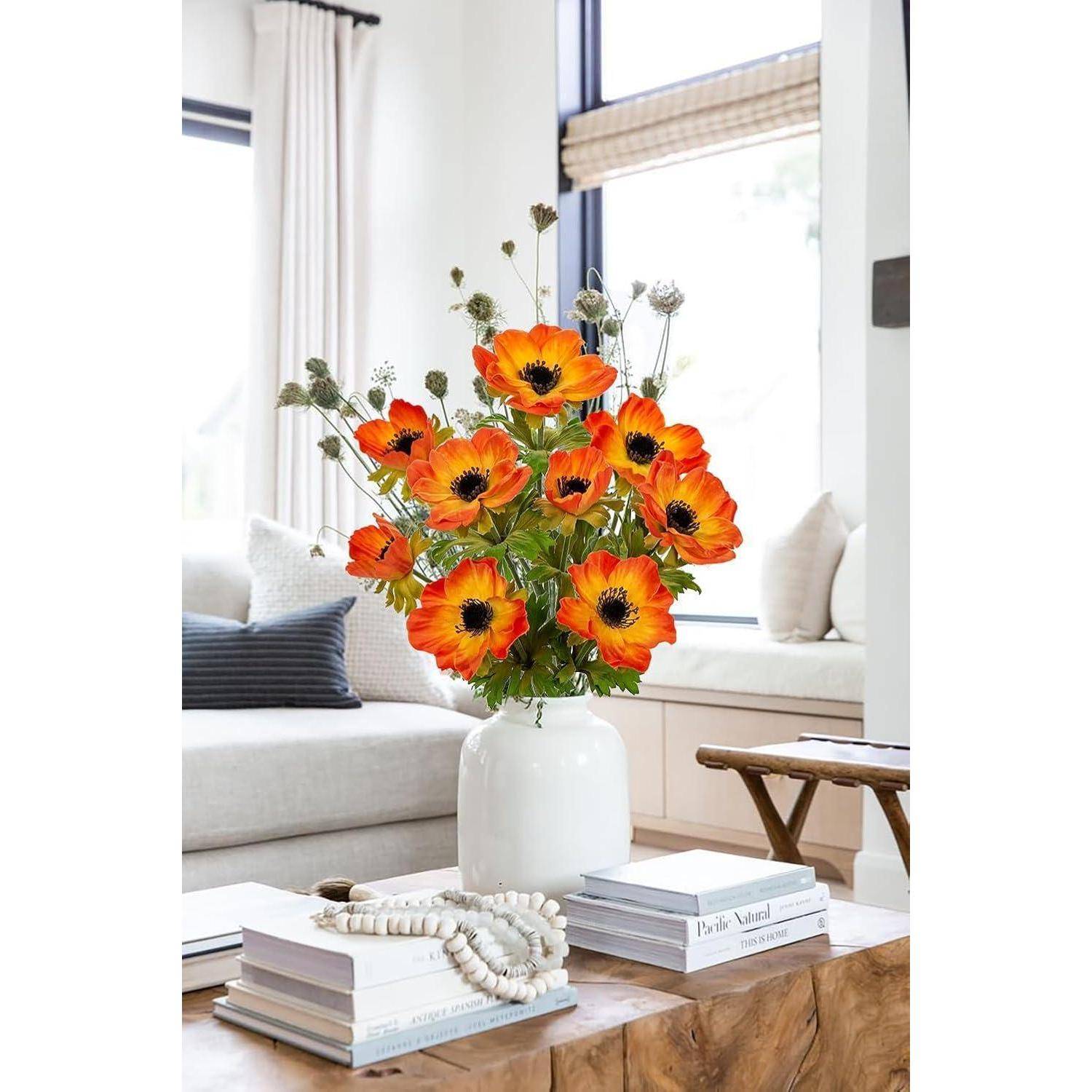 Eine weiße Vase mit lebhaft orangefarbenen Blumen auf einem Holz-Couchtisch, umgeben von Büchern und dekorativen Perlen, in einem hellen Wohnzimmer.