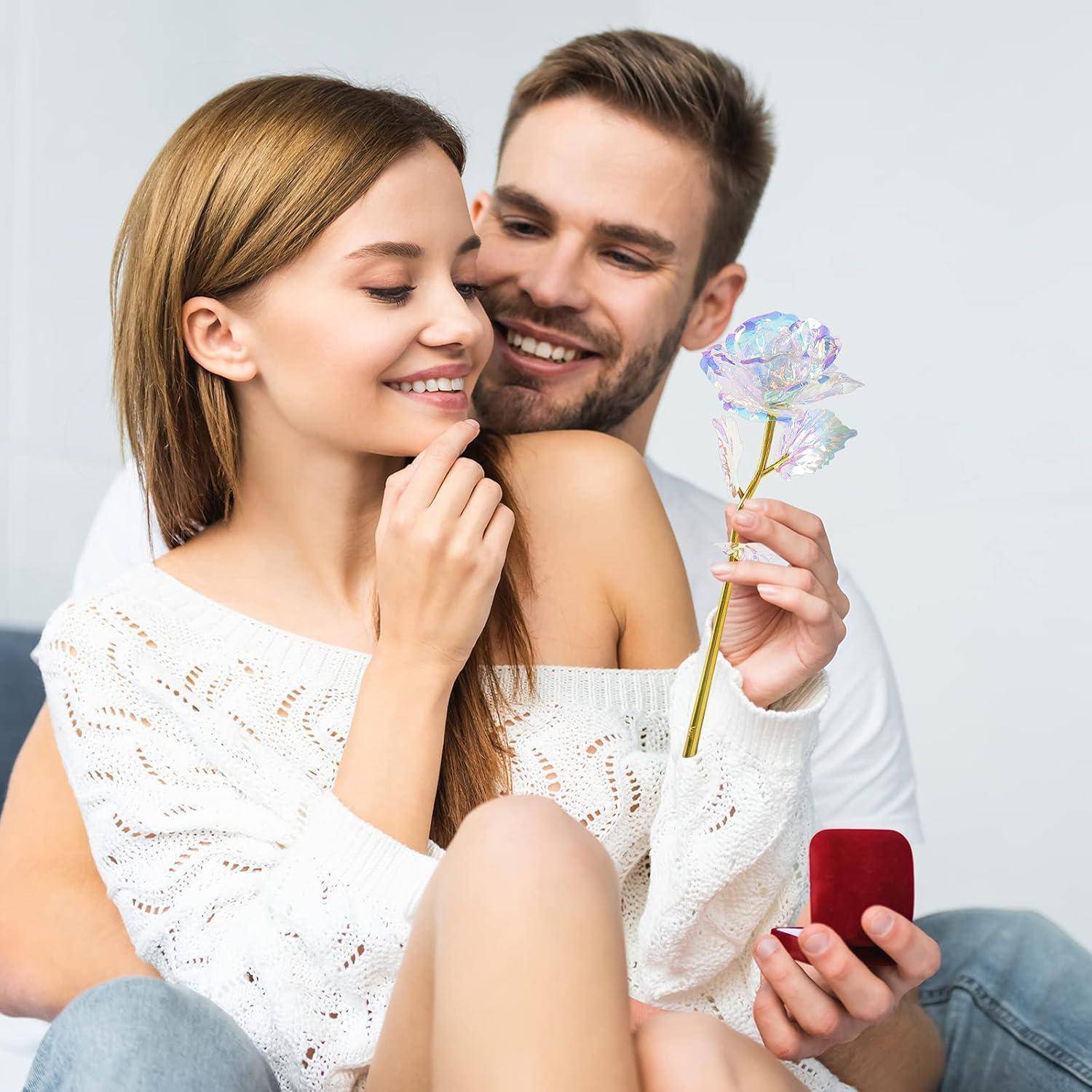 Ein Paar sitzt eng beieinander und lächelt. Der Mann hält eine transparente Blume und eine geöffnete rote Ringbox, was eine romantische Verlobungsszene andeutet.