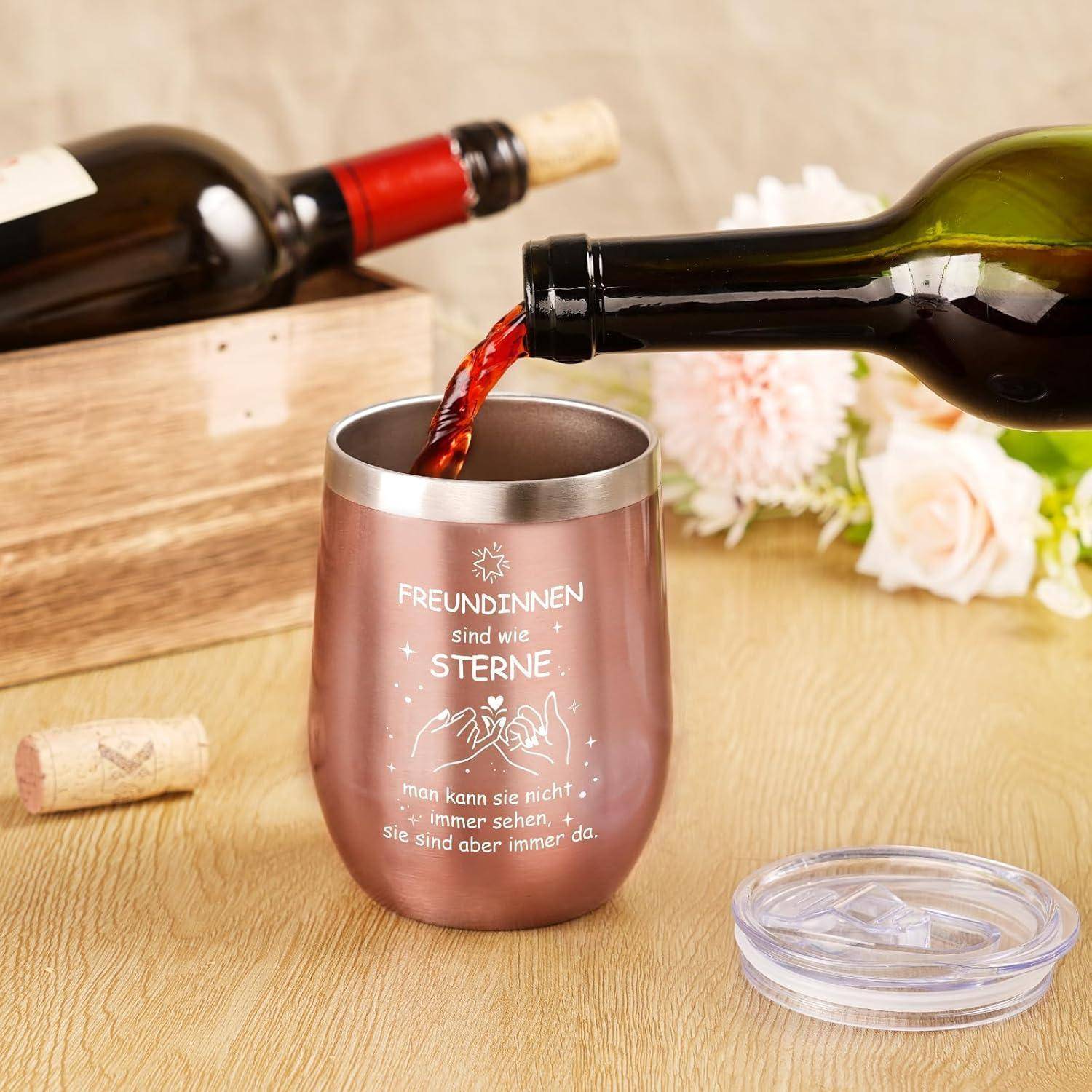 Wein wird in einen Metallbecher auf einem Holztisch gegossen, neben einem leeren Deckel und einer Weinflasche. Der Becher trägt ein deutsches Zitat über Freundschaft.