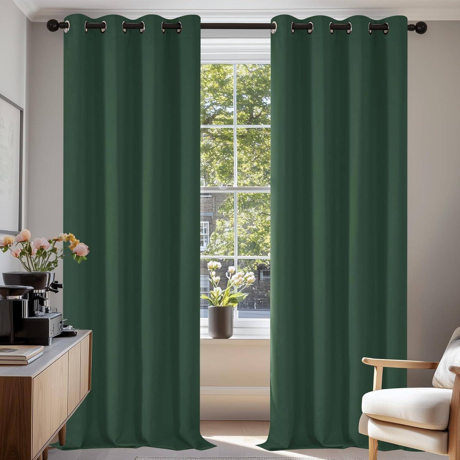 Ein Raum mit grünen Vorhängen an einem großen Fenster, durch das natürliches Licht hereinströmt. Auf einem Tisch stehen eine Kaffeemaschine und Blumen, die eine gemütliche Atmosphäre schaffen.