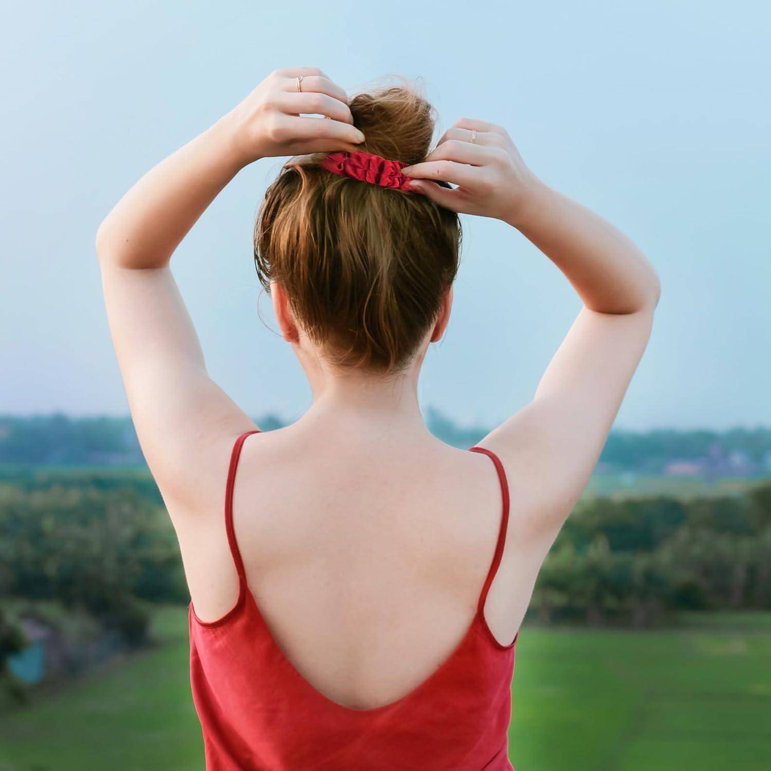 Eine Person in einem roten Oberteil steht auf einem Feld, mit dem Rücken zur Kamera, und bindet sich die Haare zu einem Dutt, vor einer Kulisse aus Bäumen und offenem Himmel.