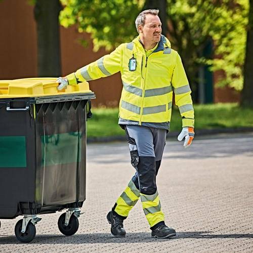 Ein Arbeiter in Warnkleidung schiebt eine große grün-gelbe Mülltonne im Freien, mit Bäumen und einem Gebäude im Hintergrund.