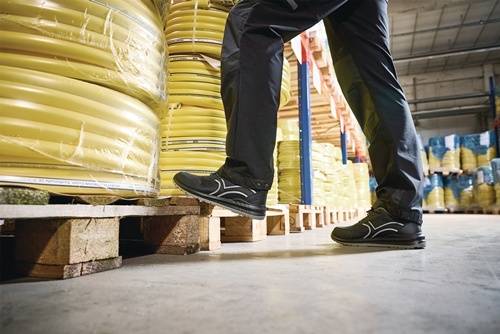 Ein Arbeiter in schwarzer Hose und schwarzen Schuhen läuft an Stapeln großer gelber Spulen auf Paletten in einer Lagerhalle vorbei.