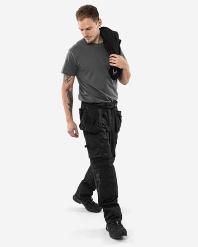 Eine Person trägt ein graues T-Shirt und schwarze Cargohose und steht, wobei sie eine Jacke über ihrer Schulter trägt.