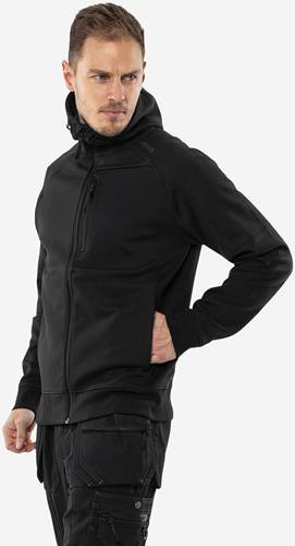 Eine Person trägt eine schwarze Sweatjacke mit Reißverschluss und hat die Hände in den Taschen, während sie vor einem einfarbigen Hintergrund steht.
