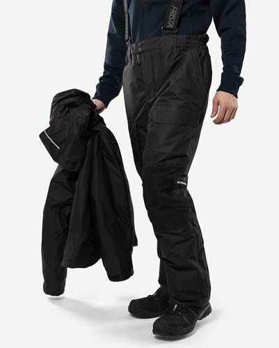 Eine Person trägt schwarze Outdoorhose und hält eine passende Jacke, bereit für kaltes Wetter. Der Fokus liegt auf der Funktionalität der Kleidung.