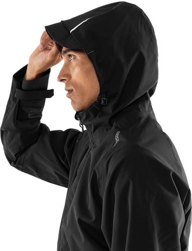 Eine Person mit einer Kapuzenjacke, die mit einer Hand die Kapuze anpasst und zur Seite blickt. Die Jacke scheint aus einem dunklen, wetterfesten Material gefertigt zu sein.