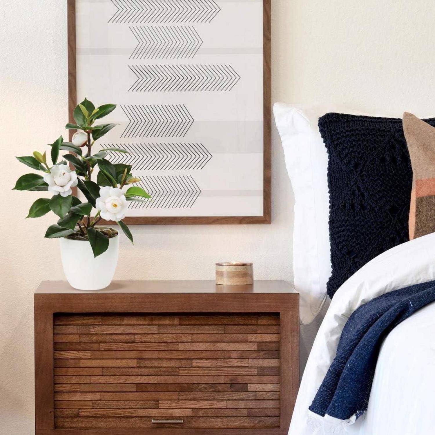 Ein Holznachttisch mit einer Topfpflanze und gerahmter geometrischer Kunstwerk darüber, neben einem Bett mit weißer und dunkelblauer Bettwäsche.