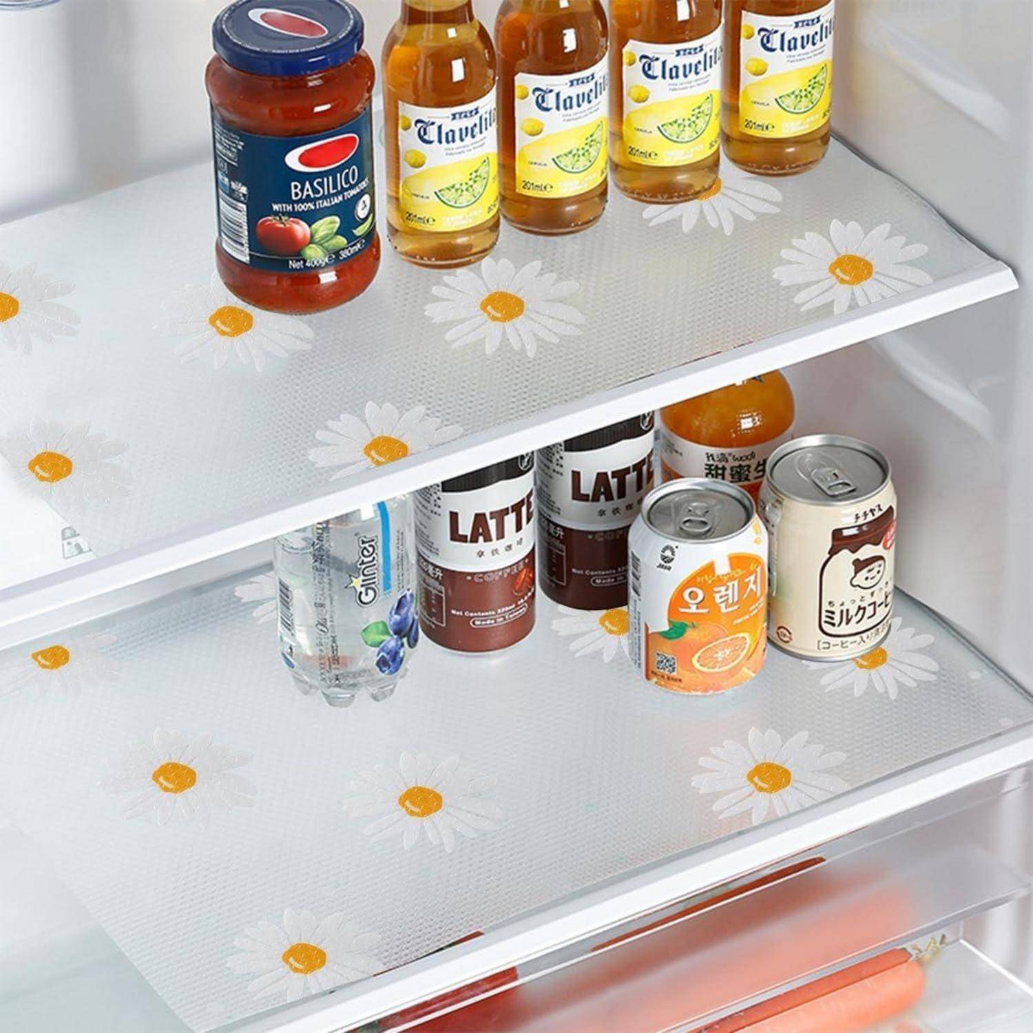 Ein offener Kühlschrank mit drei Regalen, der eine Dose Basilikosauce, Flaschen Saft, eine Wasserflasche, verschiedene Dosiergetränke und Regaleinsätze mit Gänseblümchenmuster enthält.