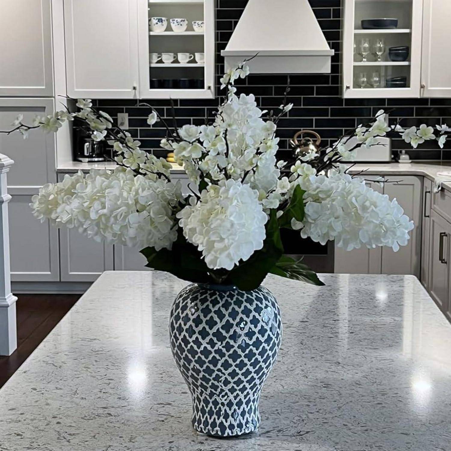 Eine dekorative Blumengestecke mit weißen Blüten in einer gemusterten Vase steht auf einer Kücheninsel, umgeben von modernen Einbauschränken und Haushaltsgeräten.