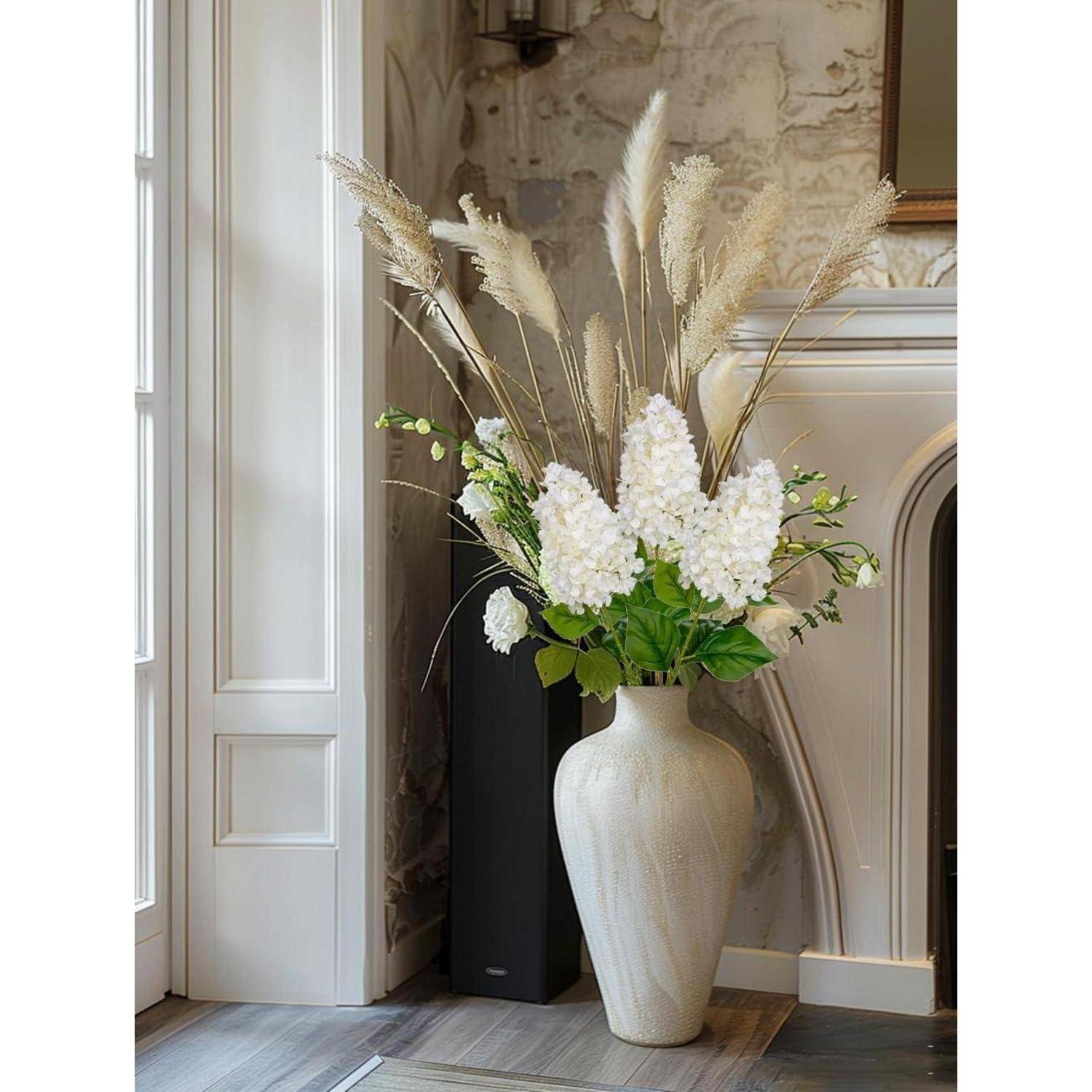 Eine hohe Vase mit weißen Blumen und Pampasgras auf einem Holzboden neben einem Fenster und einem Kamin, die eine gemütliche, elegante Raumgestaltung unterstreichen.