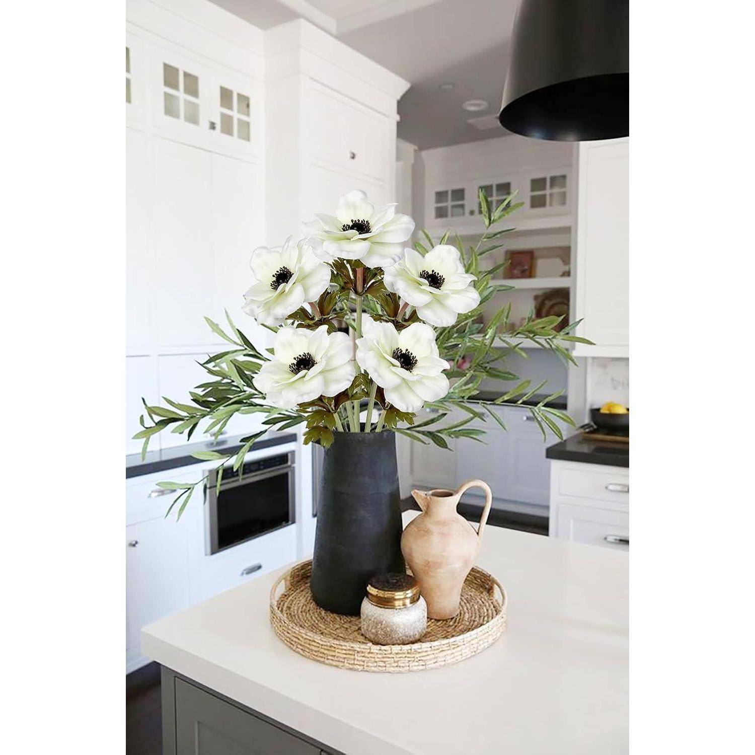 Eine dunkle Vase mit weißen Blumen und grünen Blättern auf einer Küchenarbeitsplatte, neben einem kleinen Glas und einem rustikalen Krug, in einer modernen Küche.