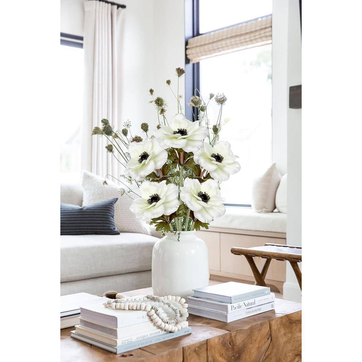 Eine weiße Vase mit weißen Blumen steht auf einem Holztisch, umgeben von Büchern und Perlen. Ein Sofa und große Fenster befinden sich im Hintergrund.