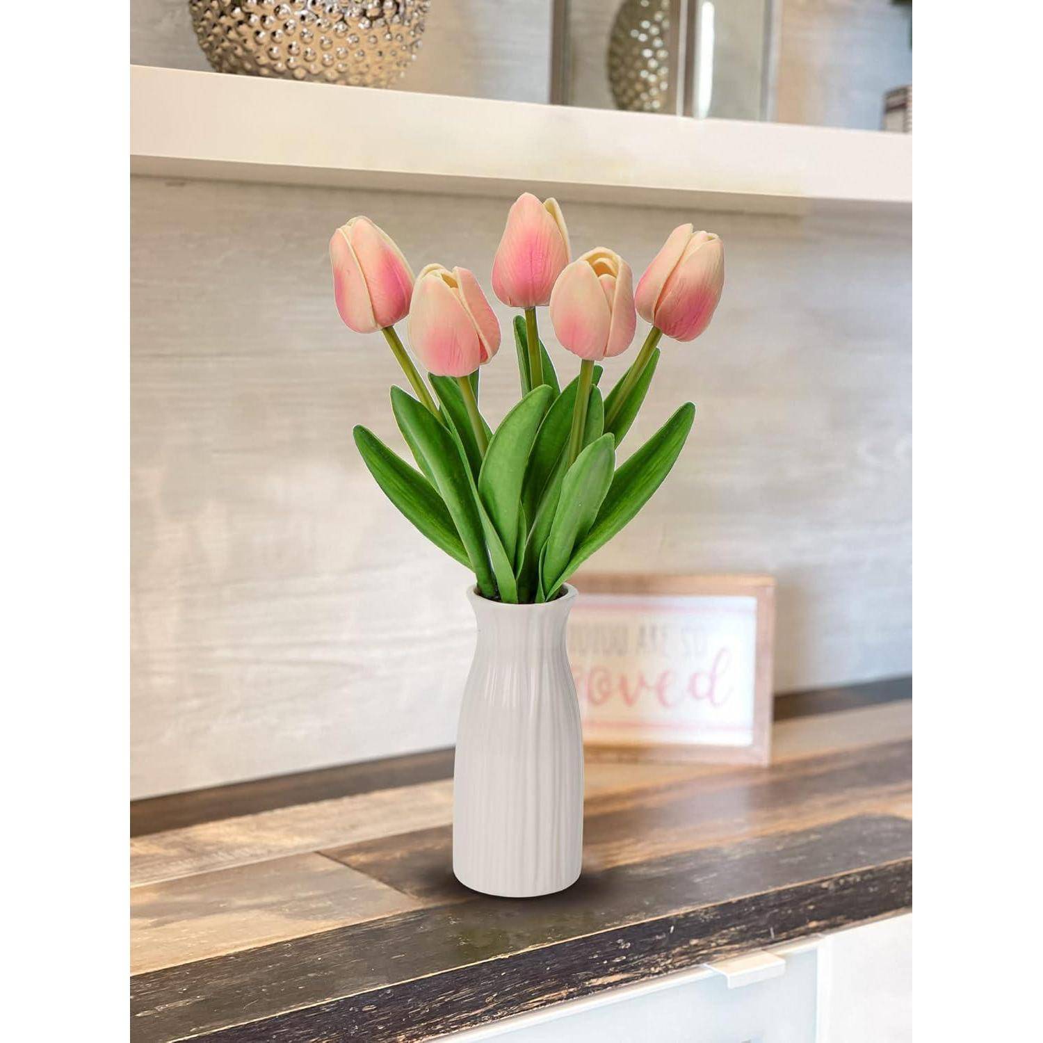 Eine weiße Vase trägt pinke Tulpen mit grünen Blättern, die auf einem Holztresen platziert sind.