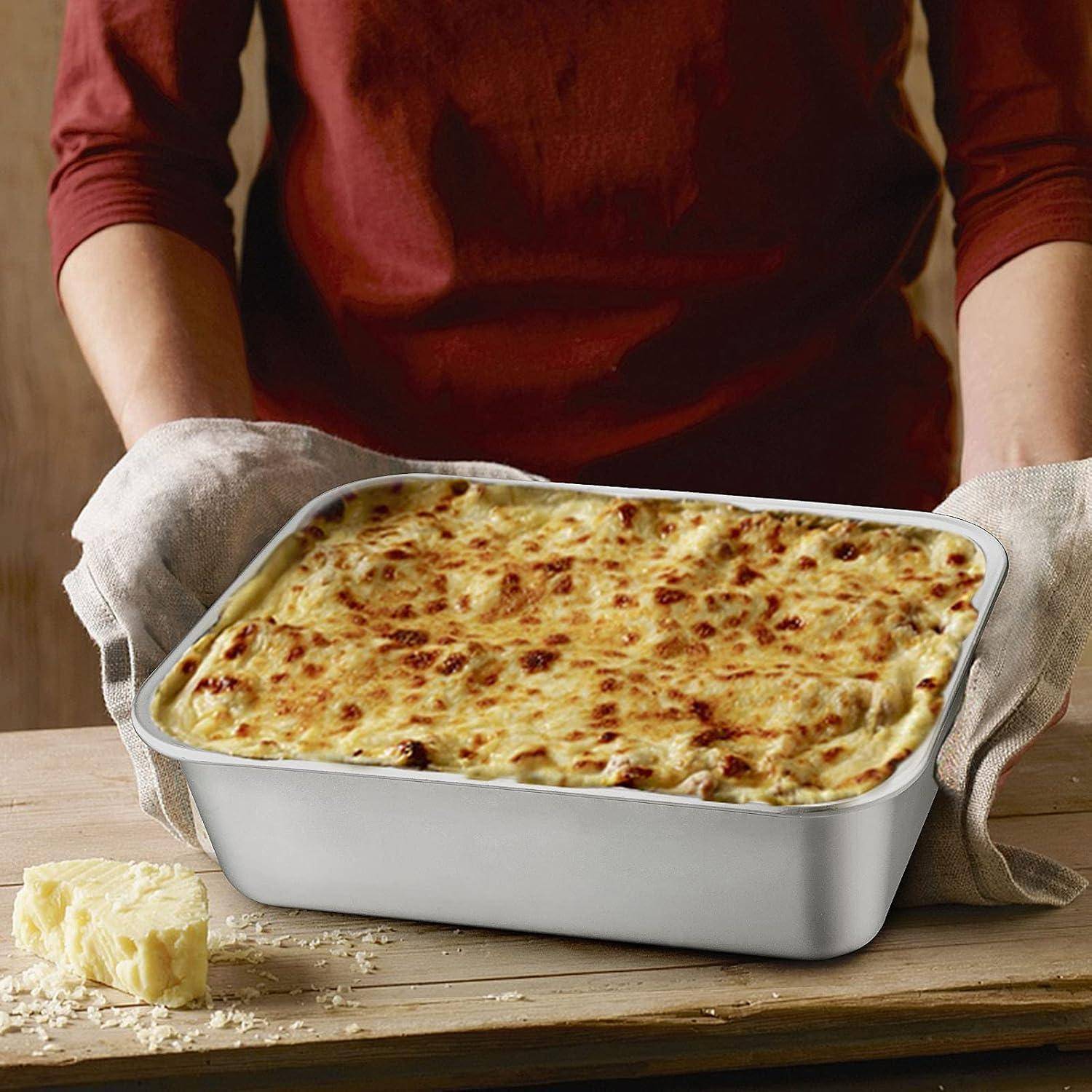 Person hält eine Pfanne mit gebackener Lasagne auf einer Holzoberfläche und trägt Ofenhandschuhe. Ein kleiner Käseblock befindet sich links daneben.